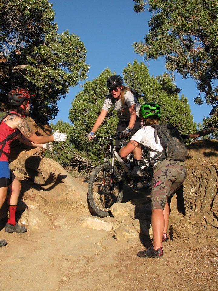 A mountain biker navigating a rocky trail while two friends offer support. One friend is gesturing with her hands, while the other is standing nearby, wearing a helmet and camo shorts, showing encouragement. The scene is set in a forested area with trees visible in the background.