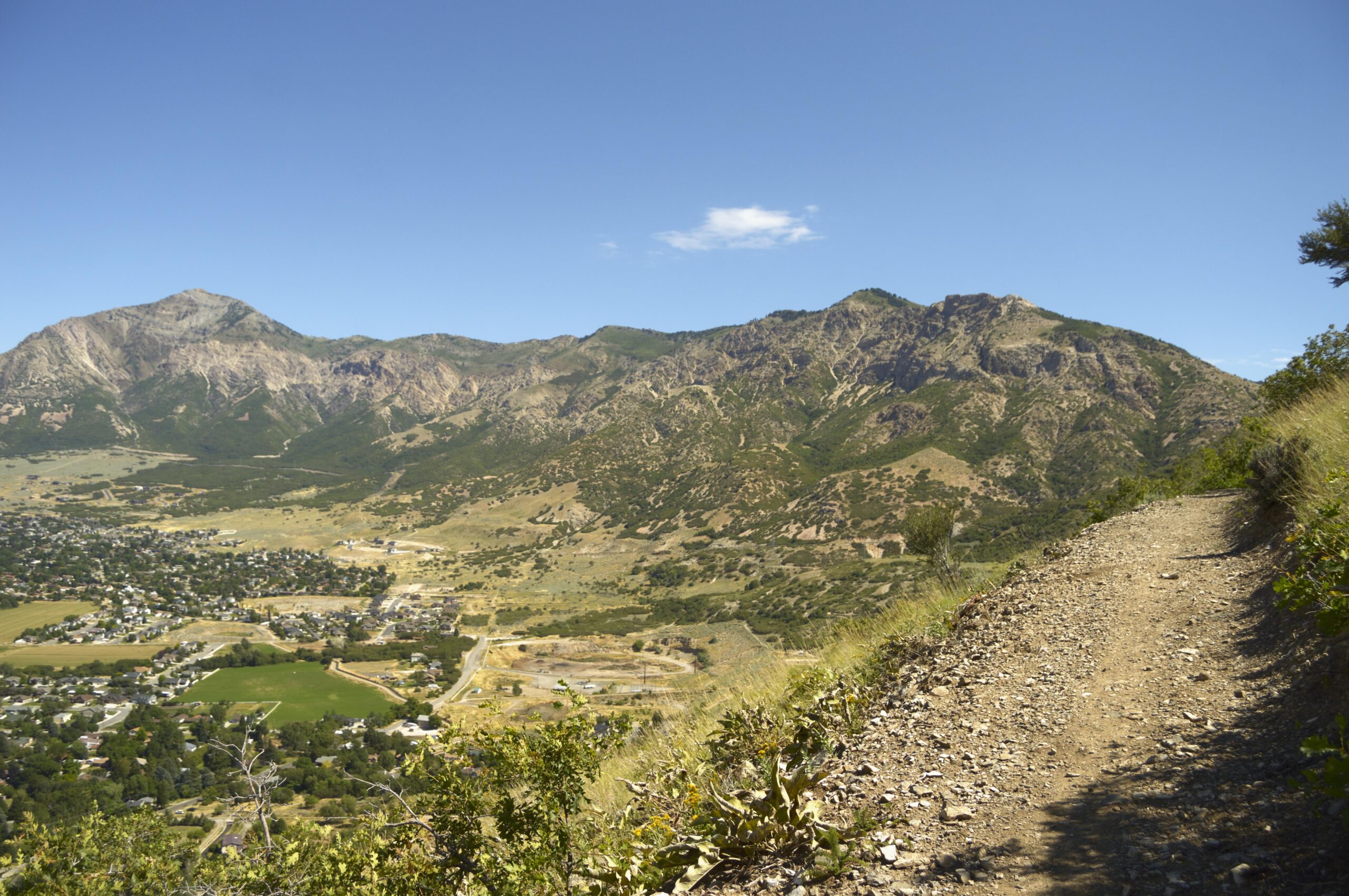 A scenic view of mountains under a clear blue sky, with a winding dirt path in the foreground and a valley with a small town visible below. The landscape features a mix of rocky terrain and patches of greenery, showcasing the natural beauty of the area. Bonneville Shoreline Ogden North Of 12th mountain bike trail.