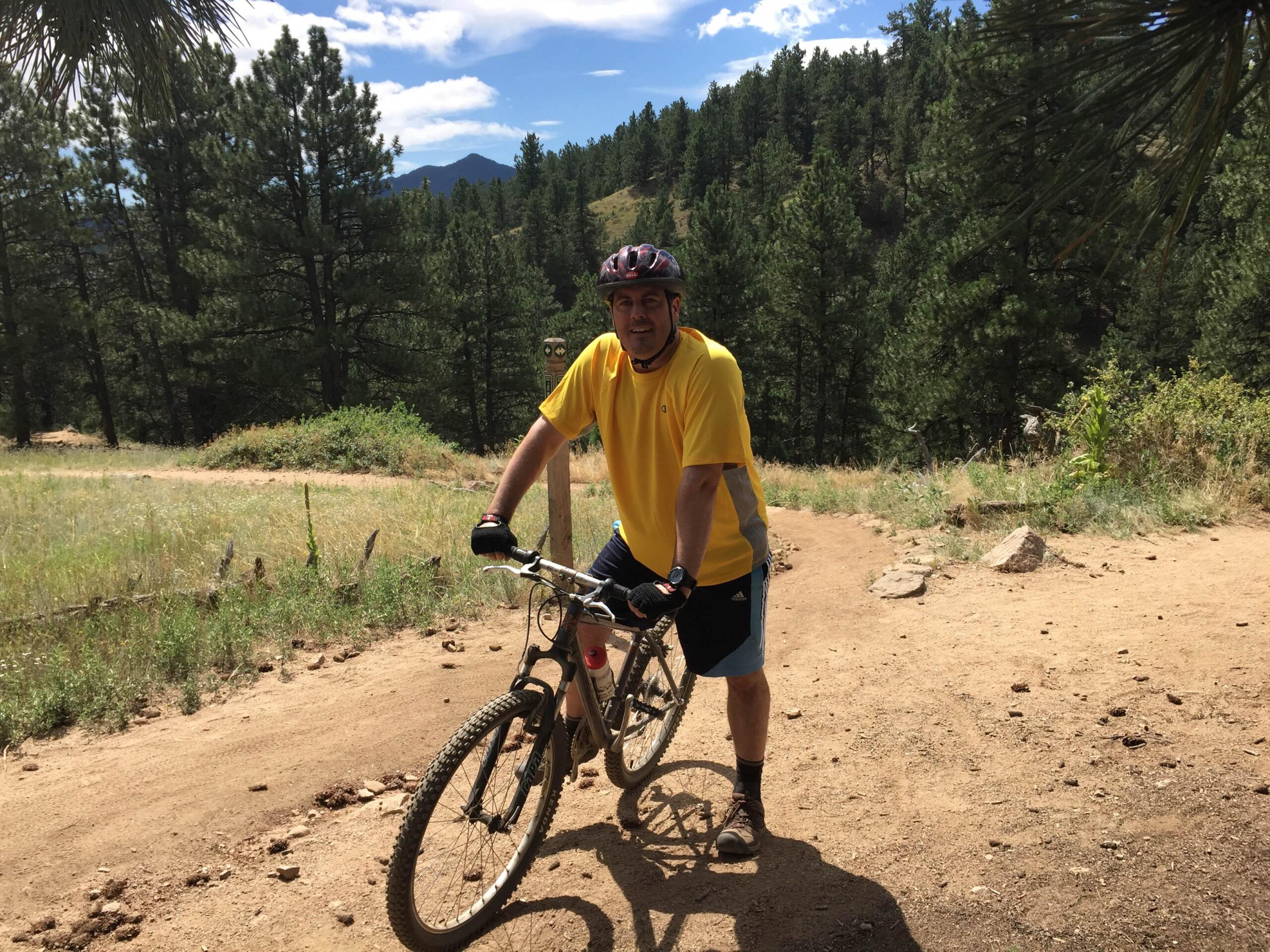 A person wearing a bright yellow shirt and shorts stands next to a mountain bike on a dirt path surrounded by tall green trees and a blue sky with clouds. The scene depicts an outdoor cycling environment, highlighting the natural landscape. Betasso Preserve mountain bike trail.