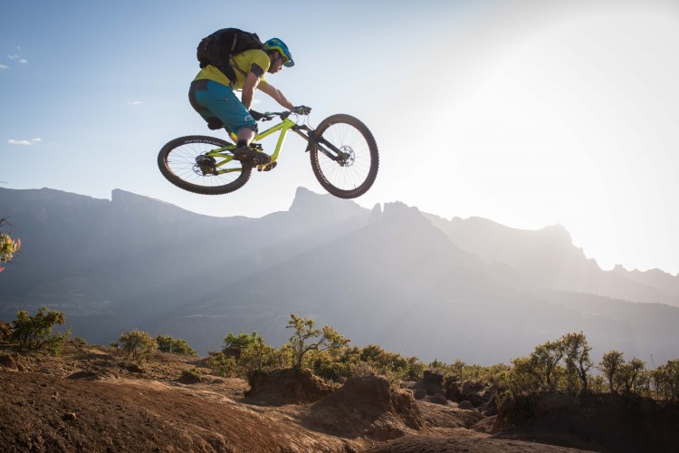 A mountain biker performing a jump over a rocky terrain, with a backdrop of mountains and a clear sky during sunset. The rider is wearing a helmet, a yellow and black shirt, and blue shorts, showcasing an athletic pose in mid-air on a bright green bike.
