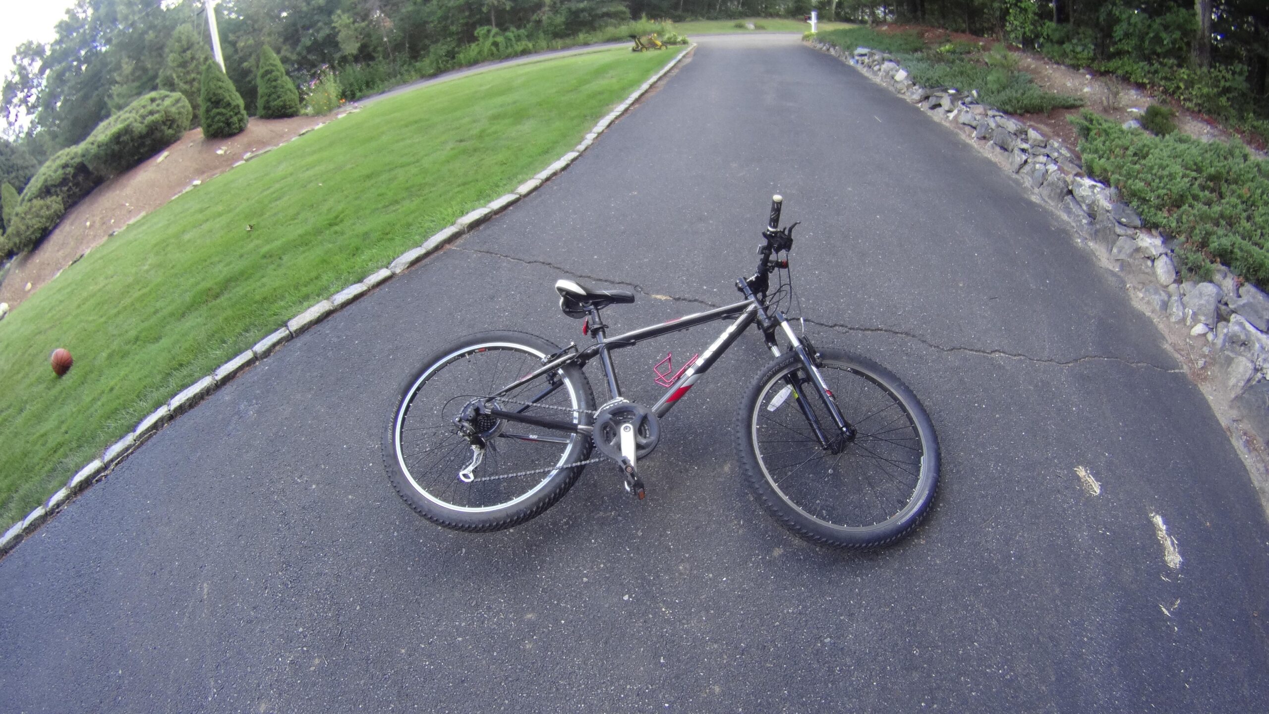 Trek 820: A black mountain bike rests on a paved driveway with gravel borders, surrounded by a lush green lawn and trees in the background. A basketball is partially visible on the grass nearby. The bike is positioned on its side, showcasing its wheels and frame.