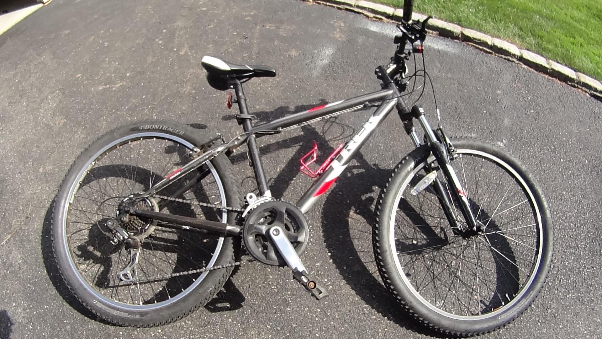 Trek 820: A black and red mountain bike parked on a paved surface, featuring thick tires, a noticeable gear system, and a water bottle holder. The bike is angled to show its frame and components clearly. Green grass and a stone border are visible in the background.