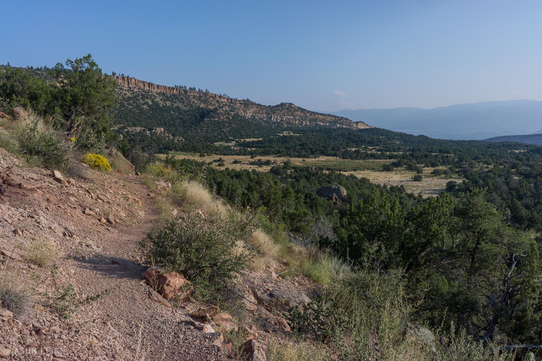 A winding dirt path leads through a scenic mountainous landscape, surrounded by green vegetation and rocky terrain. In the distance, gentle hills and cliffs stretch under a clear blue sky, creating a serene natural setting. Oil Well Flats mountain bike trail.
