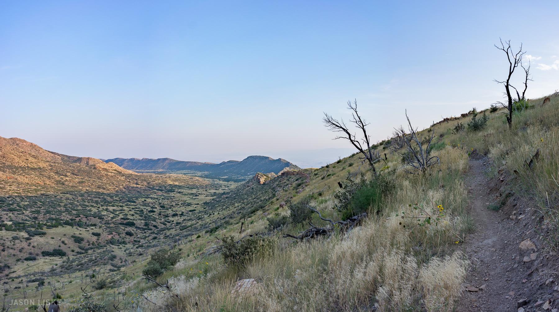 A panoramic view of a mountainous landscape featuring rolling hills covered in greenery and patches of rock. A winding dirt path leads through the foreground, bordered by tall grasses and sparse vegetation. In the distance, various mountain ridges create a layered effect under a clear blue sky. Dry tree branches are visible in the landscape, suggesting a rugged terrain. Oil Well Flats mountain bike trail.