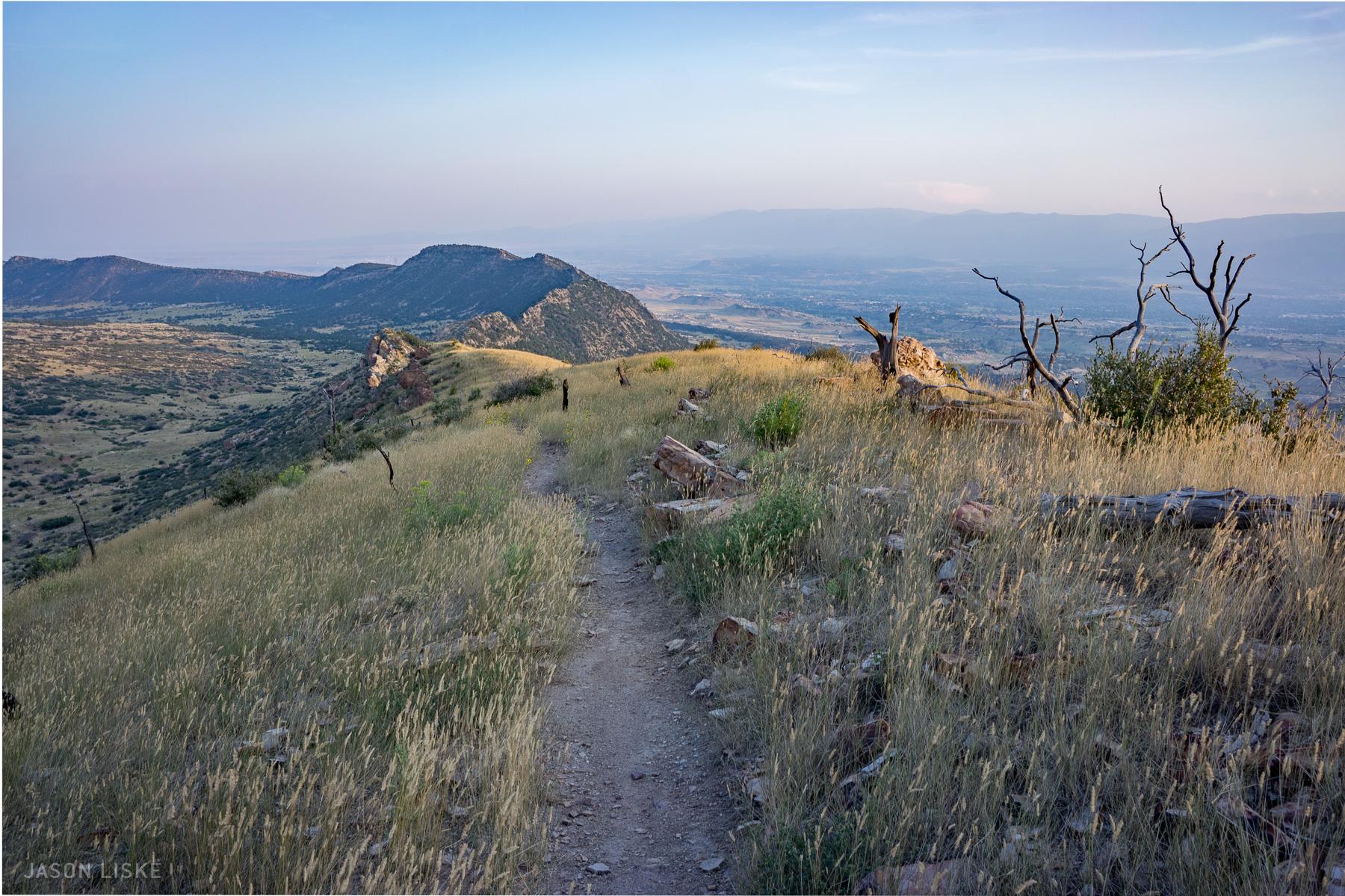 A scenic mountain landscape featuring a winding dirt path through tall grasses and rocky terrain, leading towards distant hills and valleys under a soft blue sky. Sparse trees and shrubs dot the hillside, creating a serene and natural atmosphere. Oil Well Flats mountain bike trail.