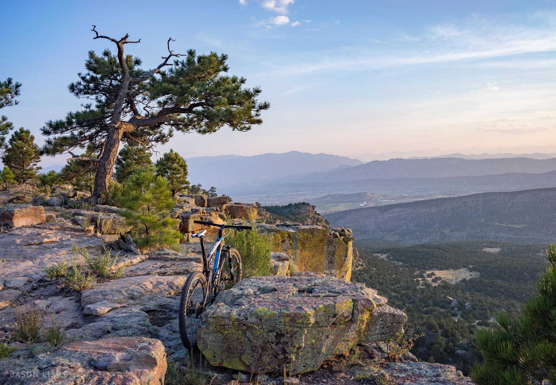 A mountain bike resting on a rocky ledge with a scenic view of valleys and mountains in the background during sunset, accompanied by trees and a clear sky. Oil Well Flats mountain bike trail.