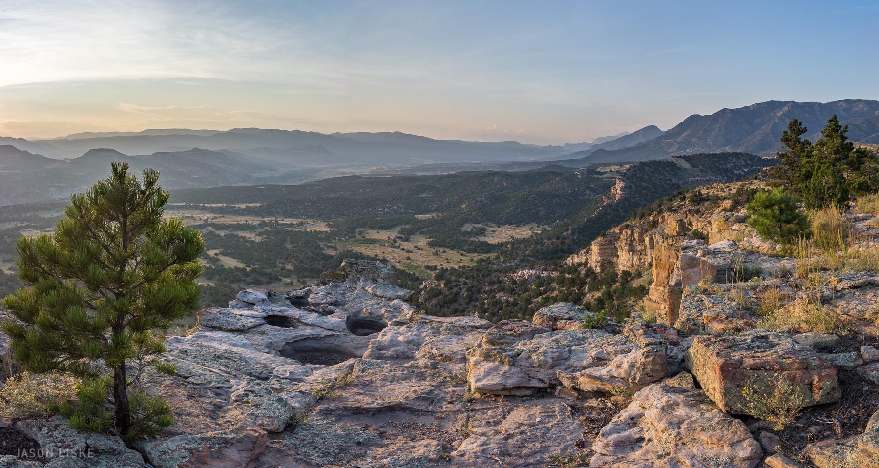 A panoramic view of a mountainous landscape during sunset, featuring rocky cliffs and a variety of trees in the foreground. The scene showcases rolling hills and valleys, with a soft, hazy atmosphere created by the setting sun illuminating the sky in warm tones. Oil Well Flats mountain bike trail.