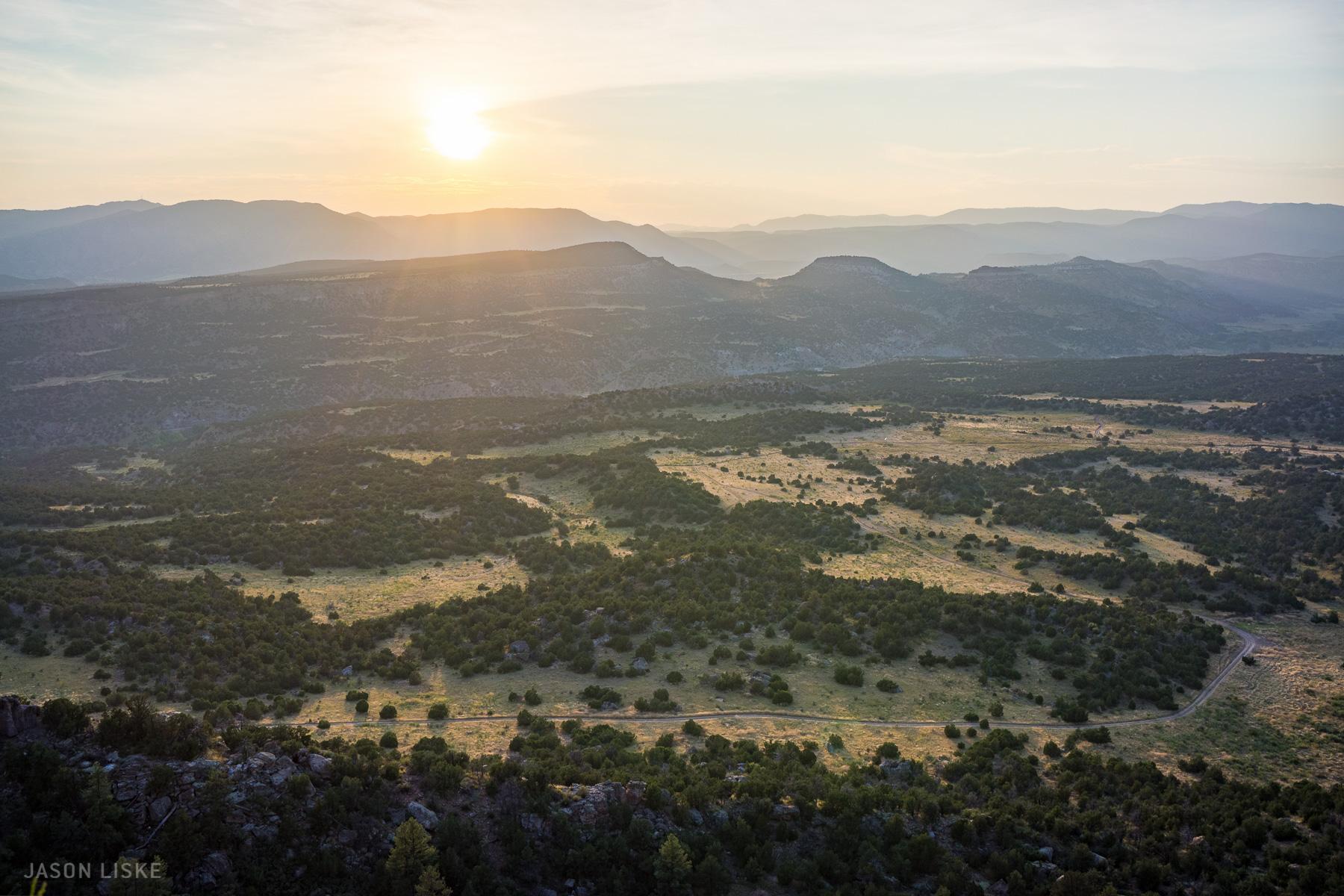 A picturesque landscape at sunrise, featuring rolling hills, a valley dotted with green shrubs, and a winding dirt road cutting through the terrain. The soft golden light of the sun casts a warm glow over the scene, highlighting the gentle contours of the hills and creating a tranquil atmosphere. Oil Well Flats mountain bike trail.