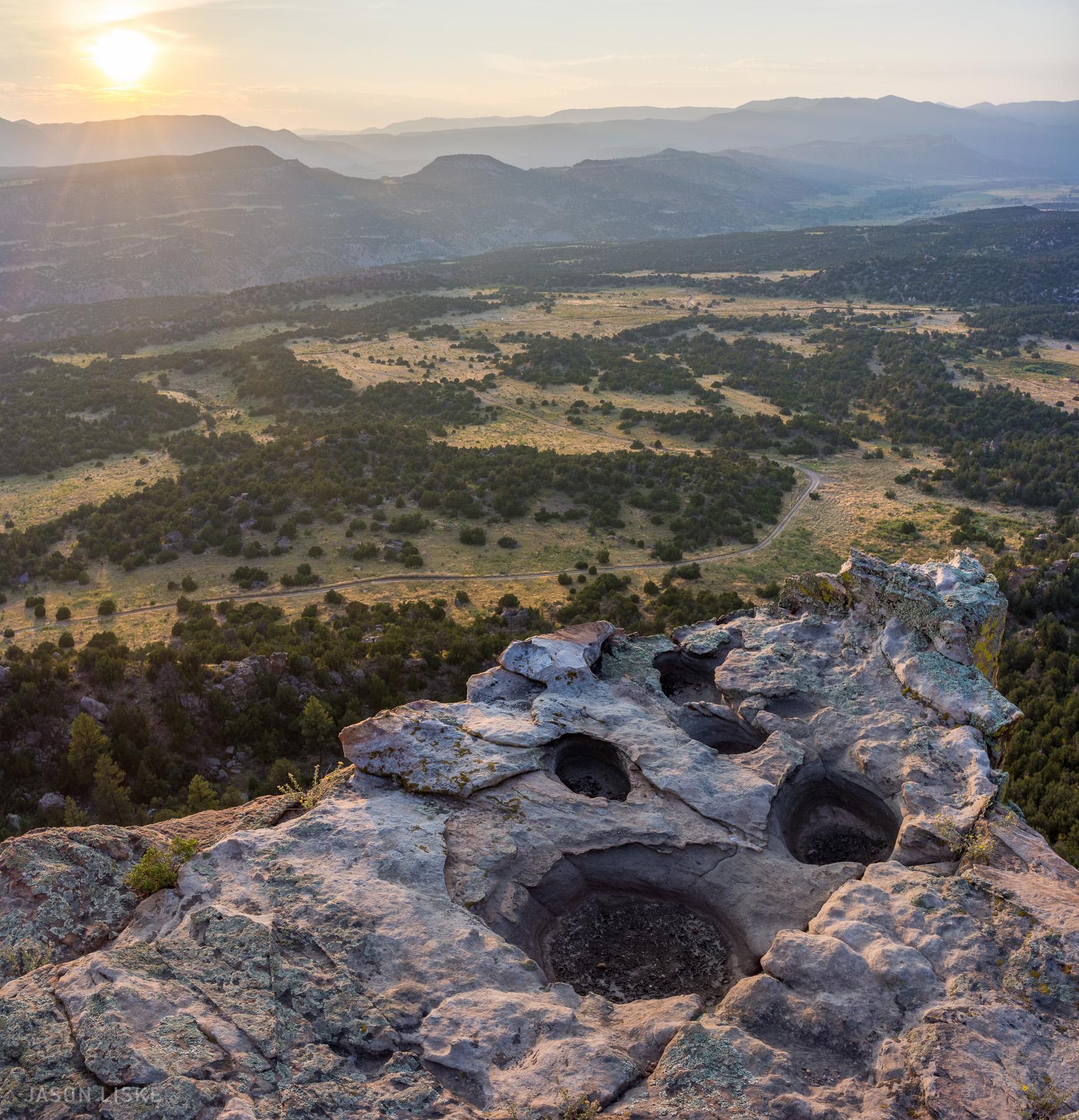 A panoramic view from the top of a rocky cliff at sunrise, showcasing the landscape of rolling hills and lush greenery below. Circular depressions are visible on the cliff's edge, and the soft light of the rising sun casts a warm glow over the scene, highlighting the rugged terrain and distant mountains. Oil Well Flats mountain bike trail.