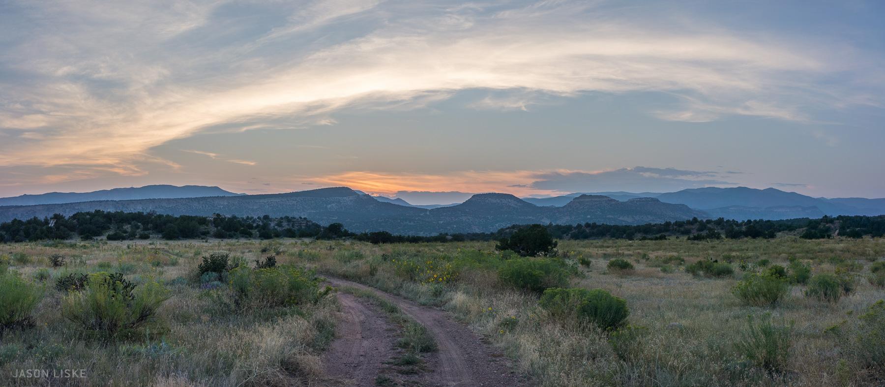 A scenic landscape featuring a winding dirt path leading through a field of tall grasses and wildflowers, set against a backdrop of rolling hills and distant mountains under a cloudy sky. The sunlight is softly illuminating the horizon, creating a tranquil and picturesque atmosphere. Oil Well Flats mountain bike trail.