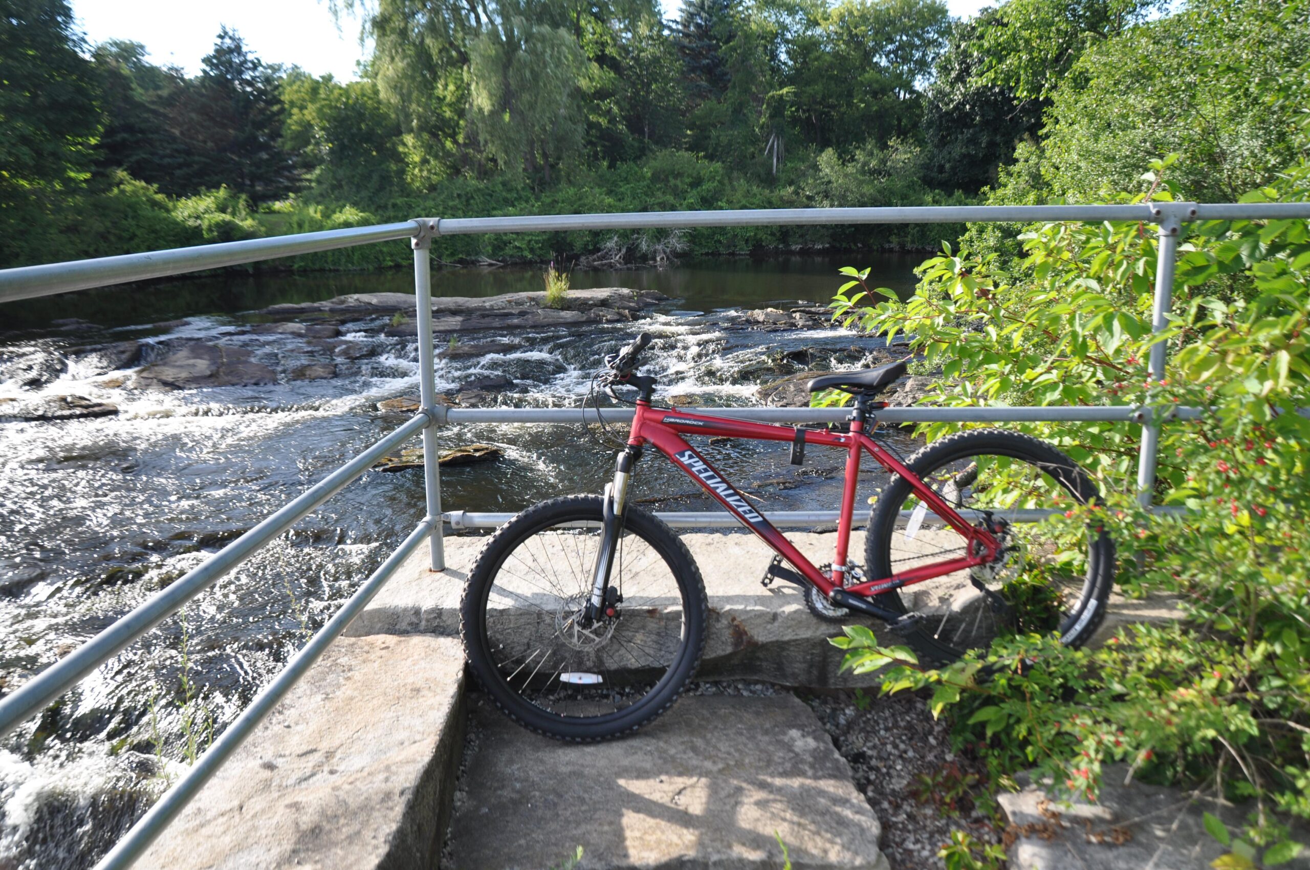 Specialized Hardrock Comp Disc: A red mountain bike rests against a railing near a rocky riverbank, with a gently flowing stream in the background surrounded by lush greenery. The scene captures a tranquil outdoor setting, perfect for cycling adventures.