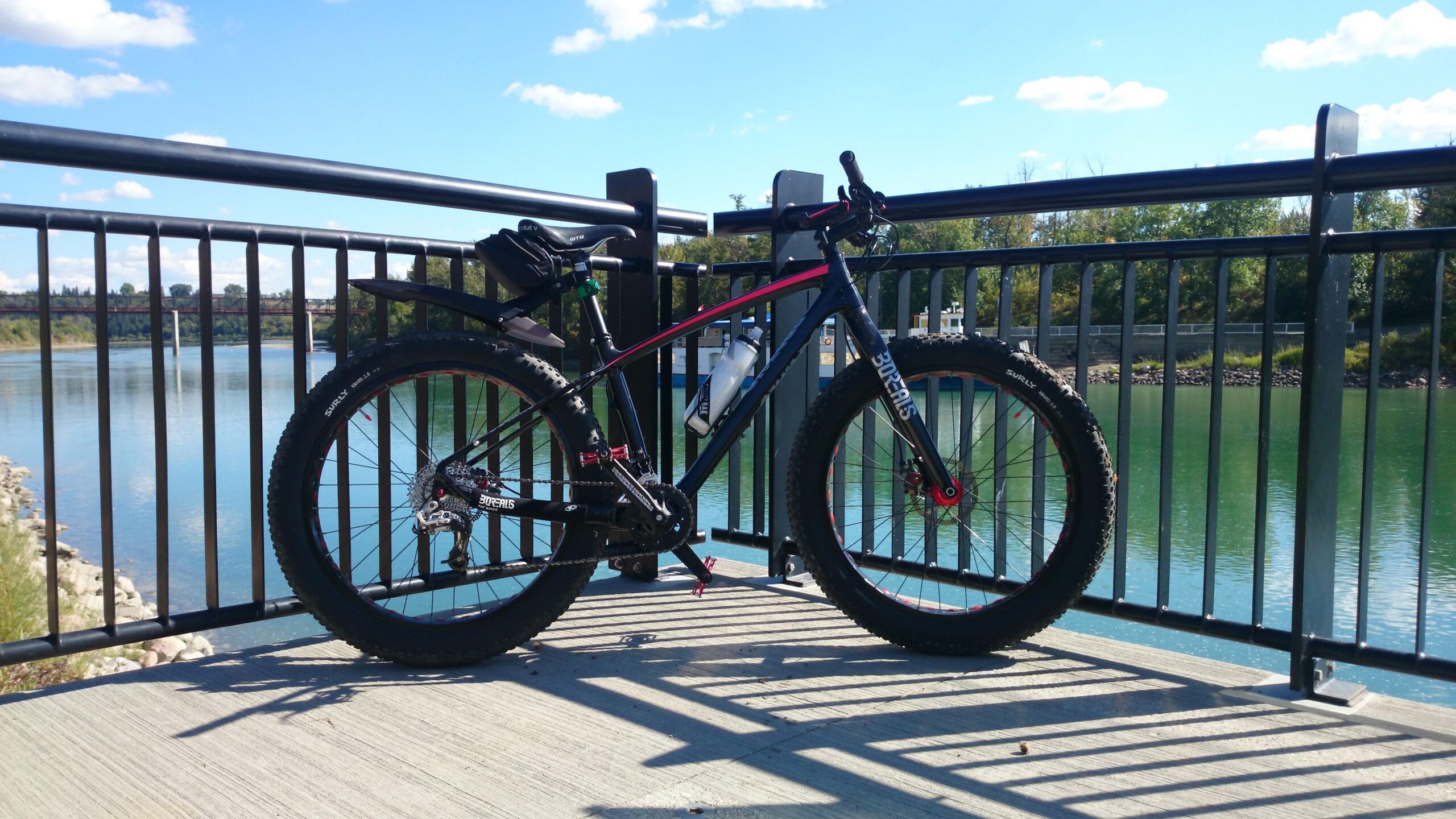 Borealis Yampa: A black and red fat bike is parked on a concrete pathway next to a railing, with a scenic view of a river and trees in the background. The bike has wide tires, a water bottle mounted on its frame, and is positioned in sunlight, casting shadows on the ground.