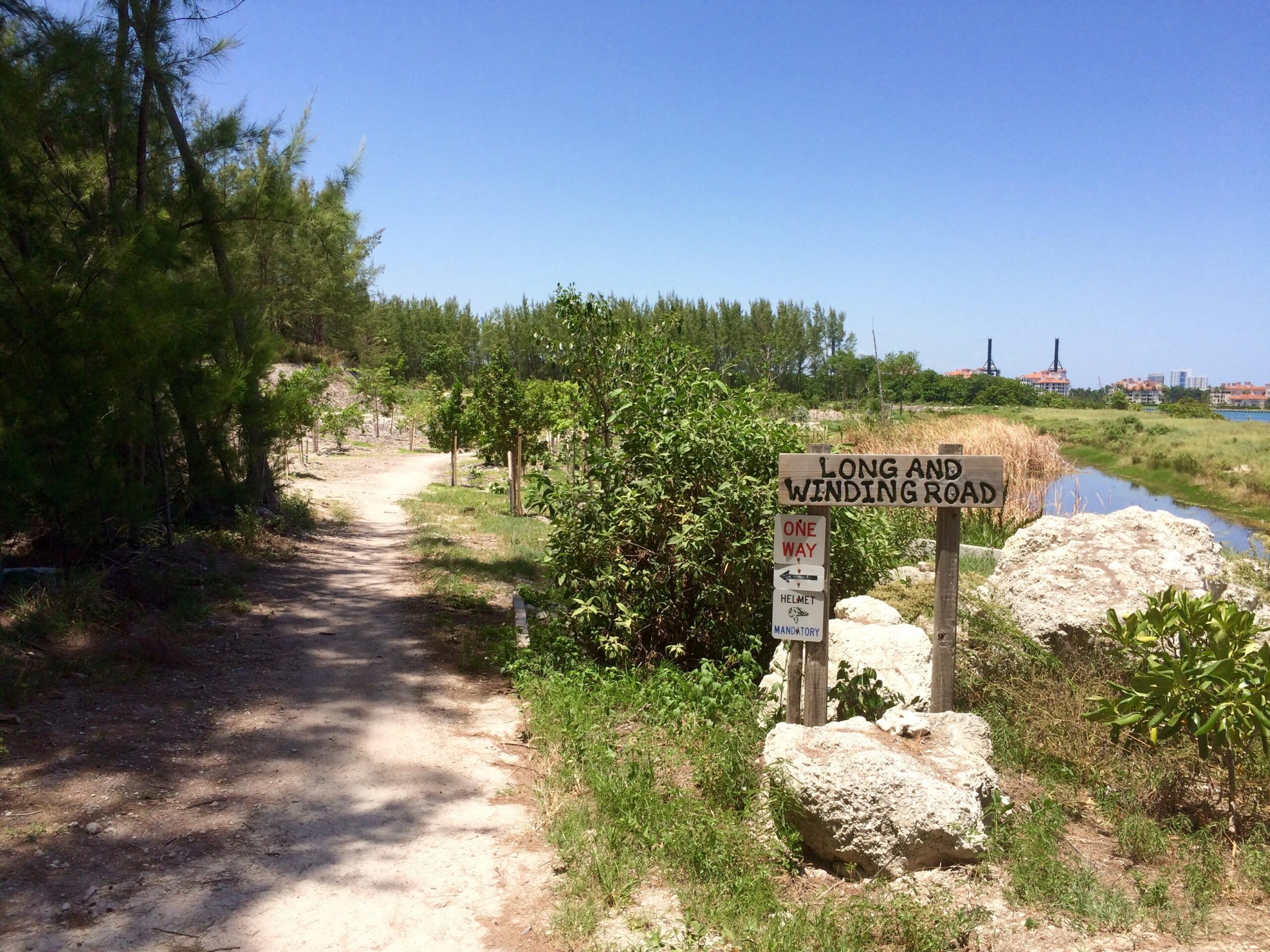 A dirt path lined with greenery leads toward a sign that reads "Long and Winding Road." The area features tall trees and a waterway beside the path, with buildings visible in the distance. The sign also includes arrows indicating directions and a note about mandatory helmet use. Virginia Key North Point mountain bike trail.