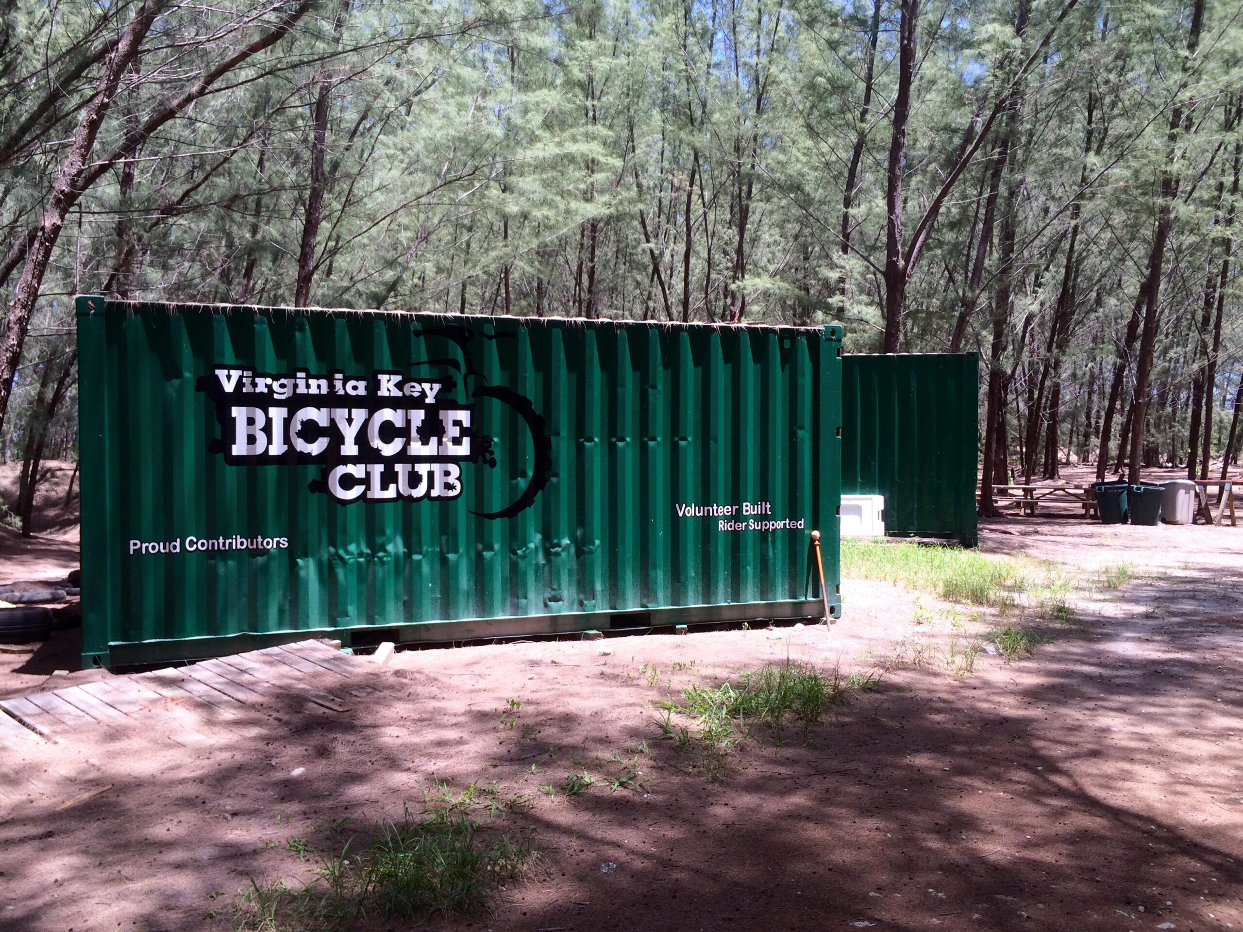 Green shipping containers marked with "Virginia Key Bicycle Club" and accompanying phrases like "Proud Contributors" and "Volunteer Built, Rider Supported" are situated in a wooded area with sandy ground and scattered greenery. Virginia Key North Point mountain bike trail.