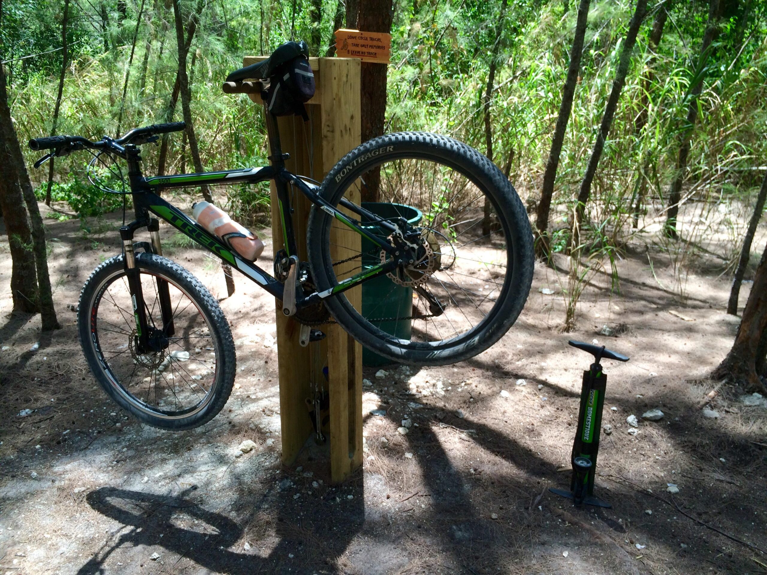 A mountain bike placed on a repair stand in a wooded area, with a bicycle pump positioned nearby. The bike has a water bottle attached to it, and trees and tall grass can be seen in the background. Virginia Key North Point mountain bike trail.