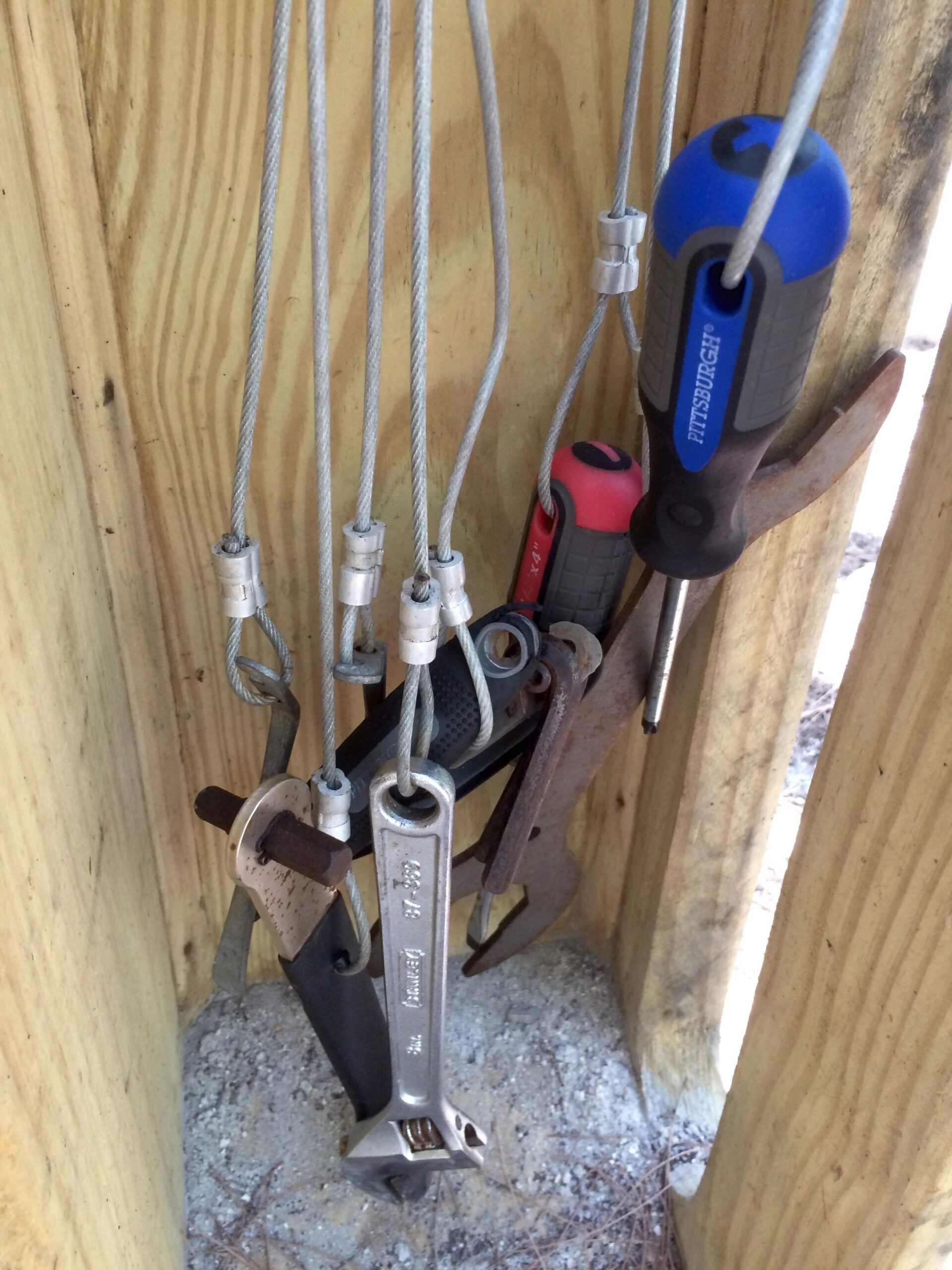 A collection of various hand tools, including adjustable wrenches and screwdrivers, hanging from cables in a wooden structure. The background features wooden planks and gravel. Virginia Key North Point mountain bike trail.