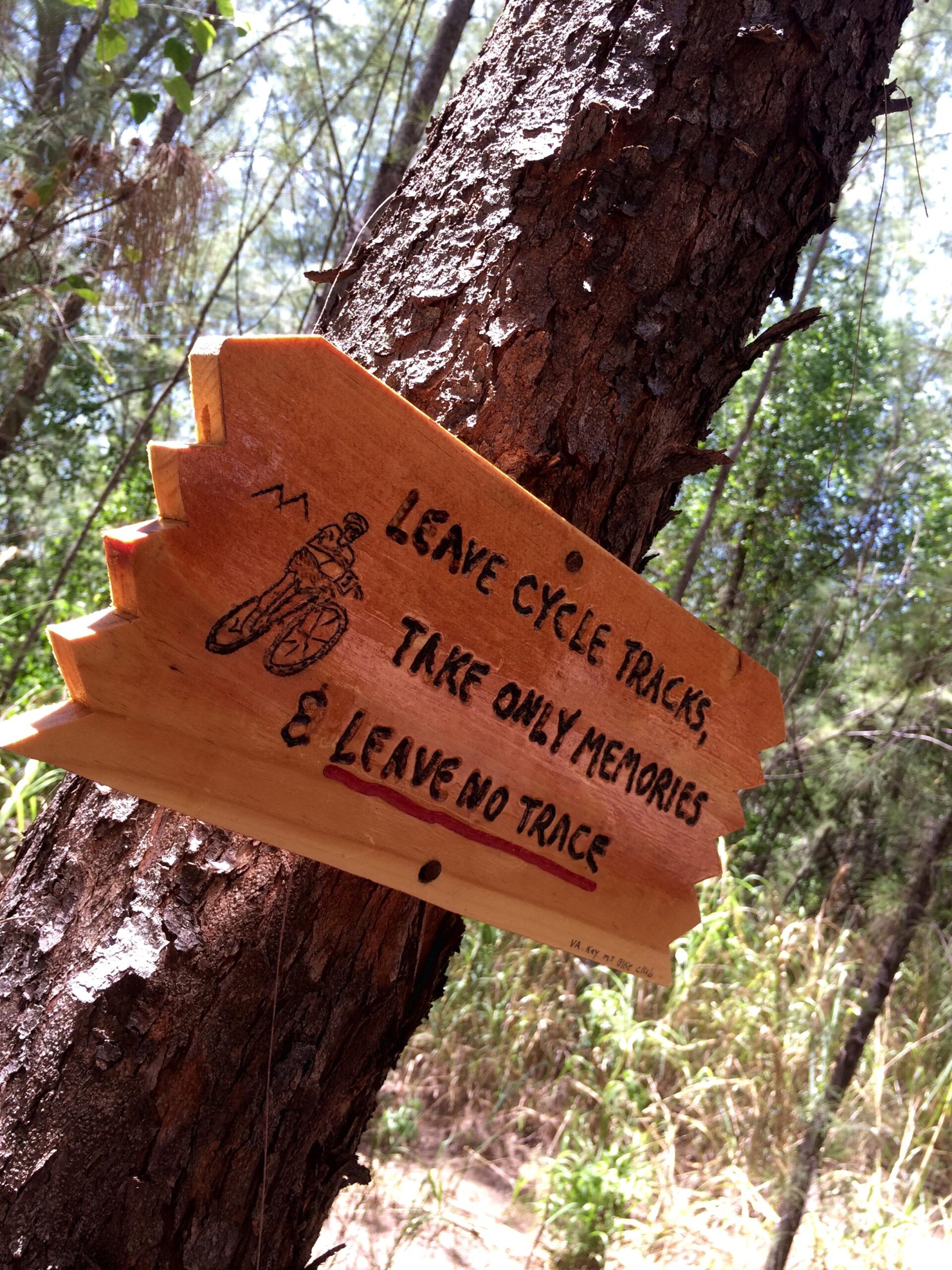 A wooden sign attached to a tree in a forest, featuring the message: "Leave cycle tracks, take only memories & leave no trace." The sign includes an illustration of a cyclist. The surrounding environment includes green foliage and a natural, rustic setting. Virginia Key North Point mountain bike trail.
