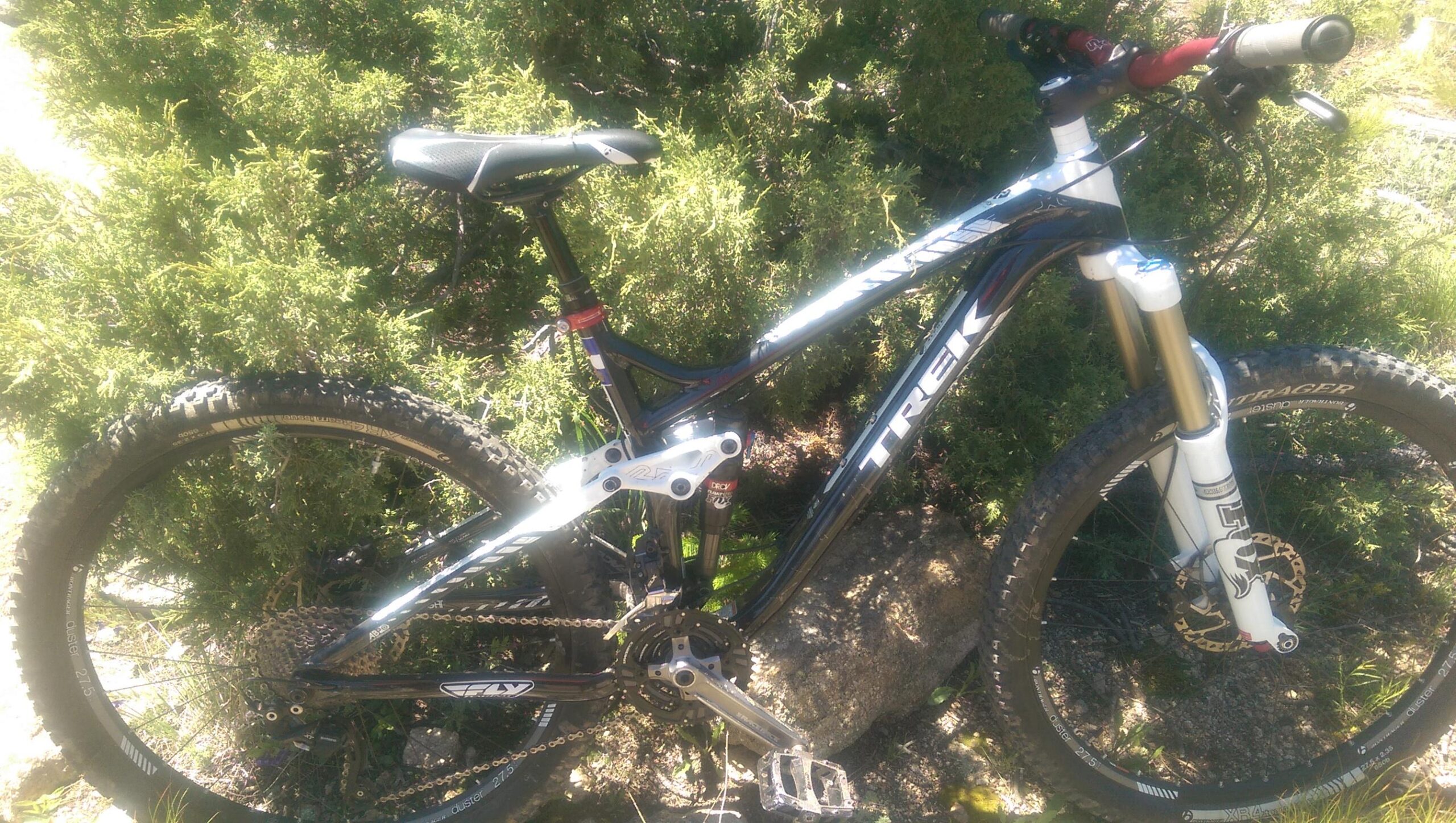 Trek Trek Remedy 8 27.5: A black and white mountain bike leaning against a bush, showcasing its suspension and handlebars. The bike features thick tires suitable for off-road terrain and has a Trek logo visible on the frame.