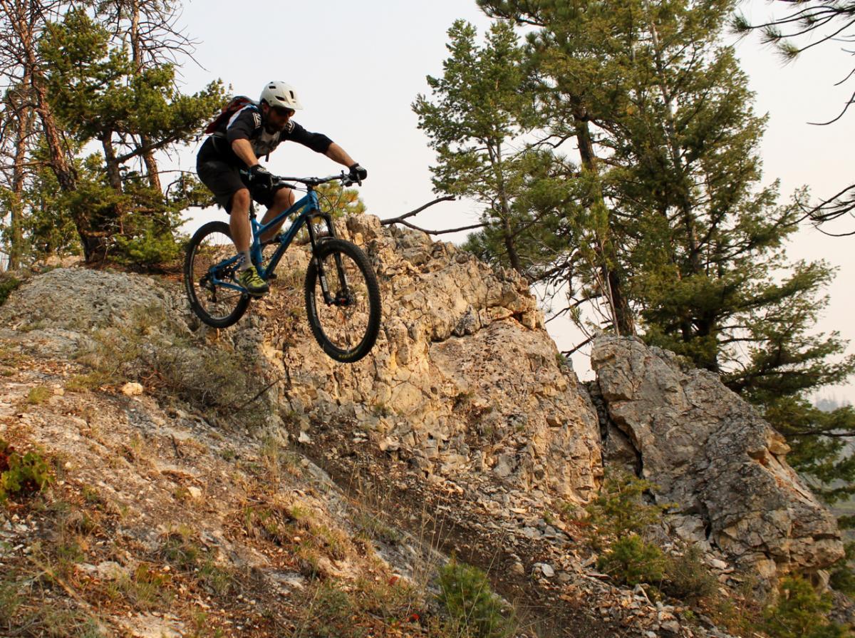 A mountain biker performing a jump over rocky terrain, surrounded by trees and a natural landscape. South Hills Trail System mountain bike trail.