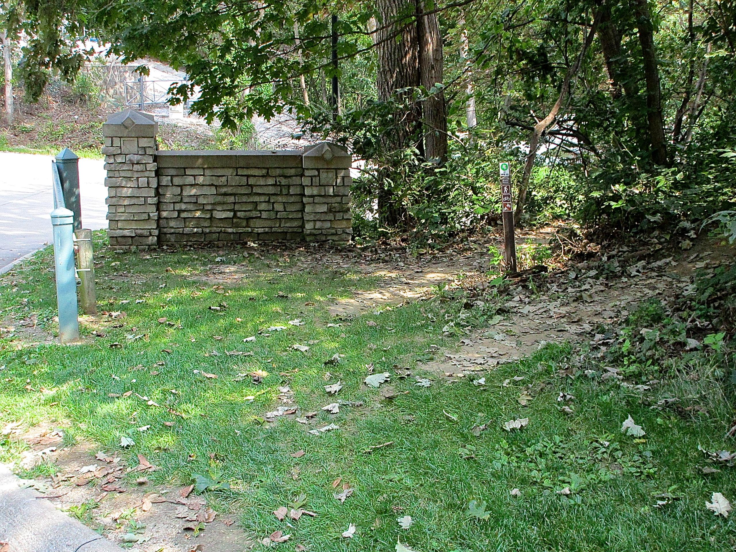 A stone wall with decorative pillars at the corners stands alongside a grassy area, featuring scattered fallen leaves. A dirt path leads into a wooded area on the right, while a street is visible in the background. A metal post is on the left, possibly indicating a trail or pathway. Lewis And Clark Monument mountain bike trail.