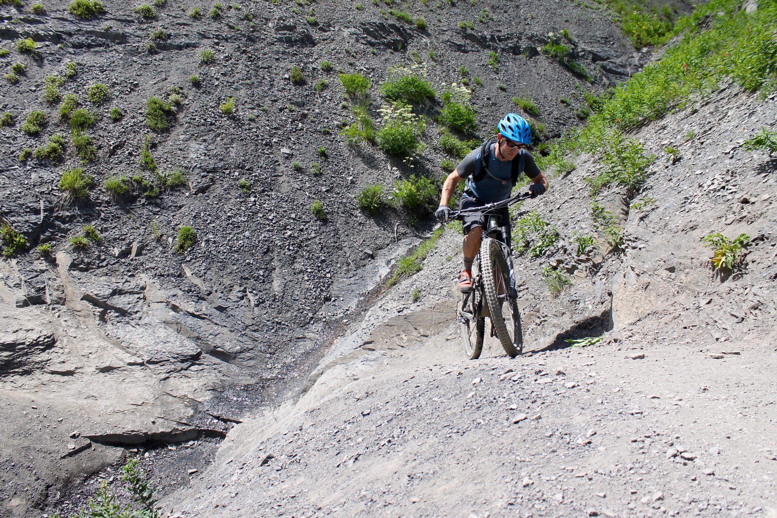 A mountain biker navigating a rocky trail surrounded by sparse vegetation. The cyclist wears a blue helmet and a backpack, pedaling uphill on a dirt path with large stones and gravel. Bright sunlight illuminates the scene, highlighting the rugged terrain. Trail 401 mountain bike trail.