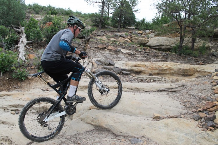 A person riding a mountain bike on a rocky trail, navigating uphill terrain surrounded by greenery and trees. The cyclist is wearing a helmet and protective gear, demonstrating an action shot as they maneuver over rough patches of rock and dirt.