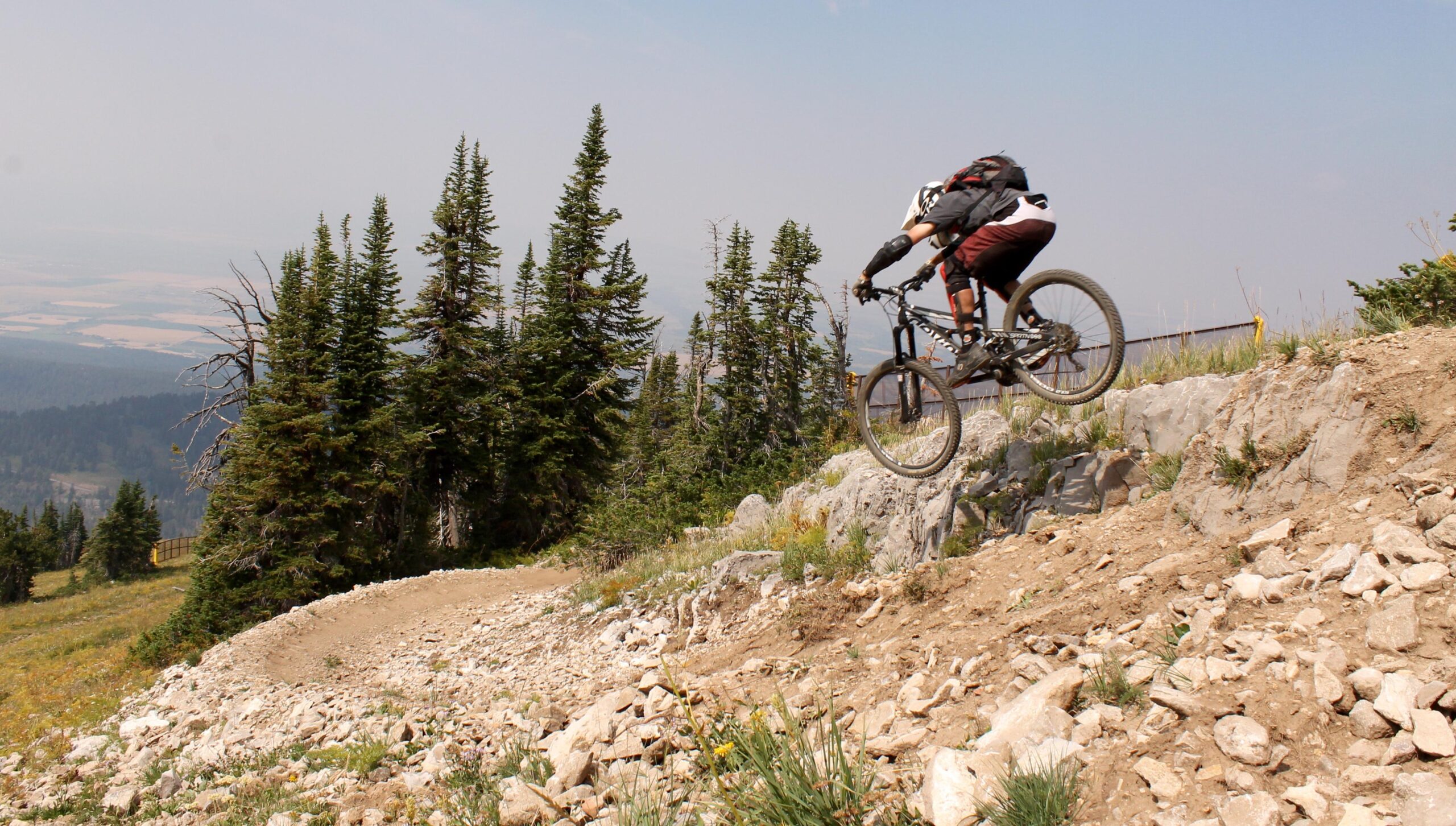 A mountain biker performing a jump over rocky terrain, surrounded by tall trees and a mountainous landscape. The biker is captured mid-air, showcasing an action-packed moment on a rugged trail. Grand Targhee Bike Park mountain bike trail.
