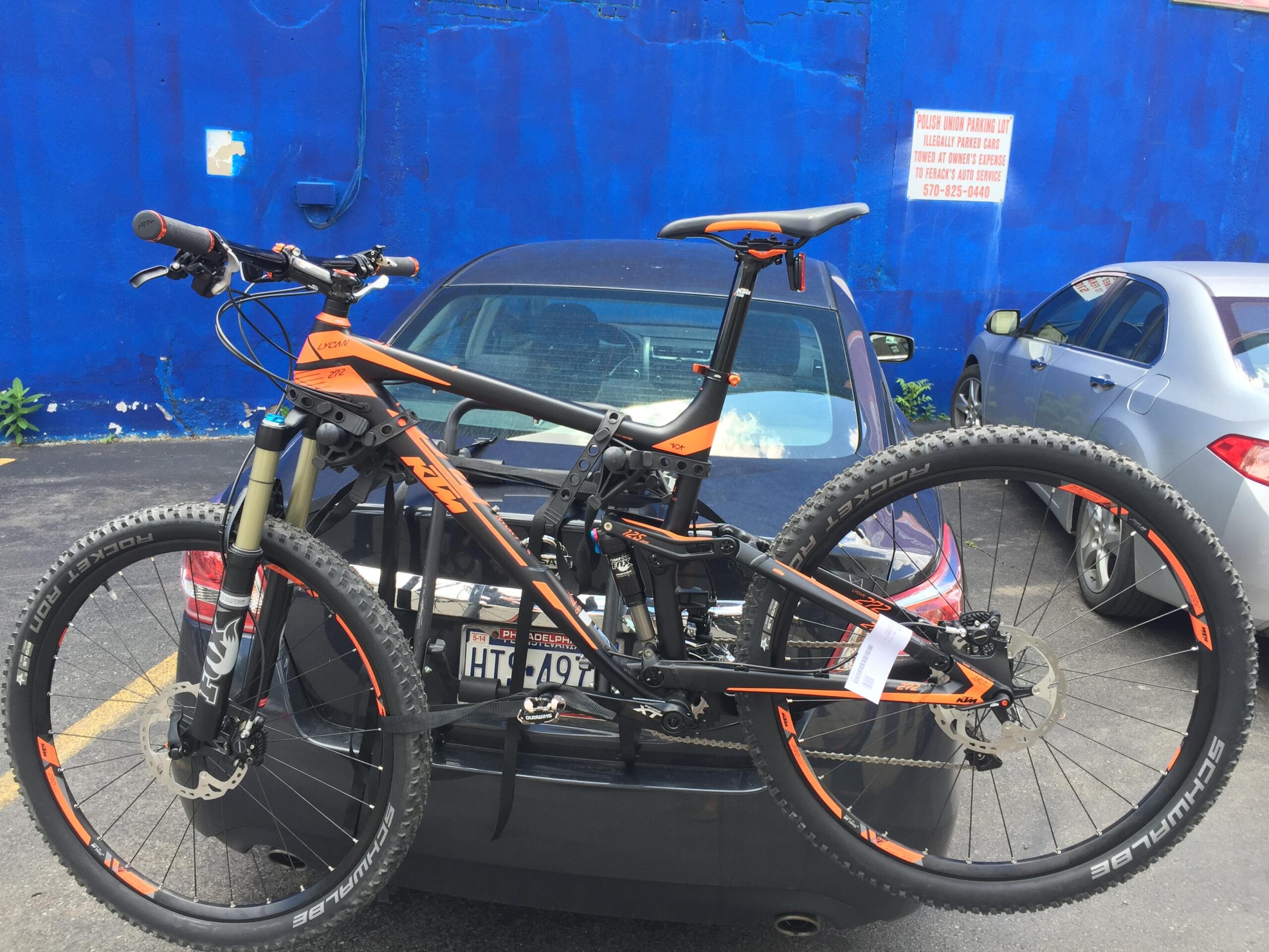 Ktm Lycan 272: A mountain bike with orange accents is secured on the back of a car parked in a lot. In the background, a bright blue wall and a sign about parking regulations are visible. The scene captures a sunny day with a clear view of the parked vehicles.