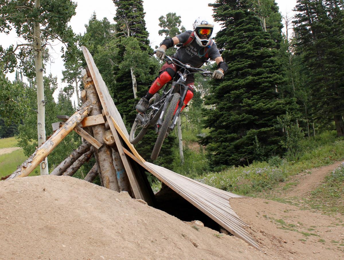 A mountain biker in protective gear jumps off a wooden ramp made from logs and planks, soaring through the air against a backdrop of green trees and a dirt path. Otter Slide mountain bike trail.