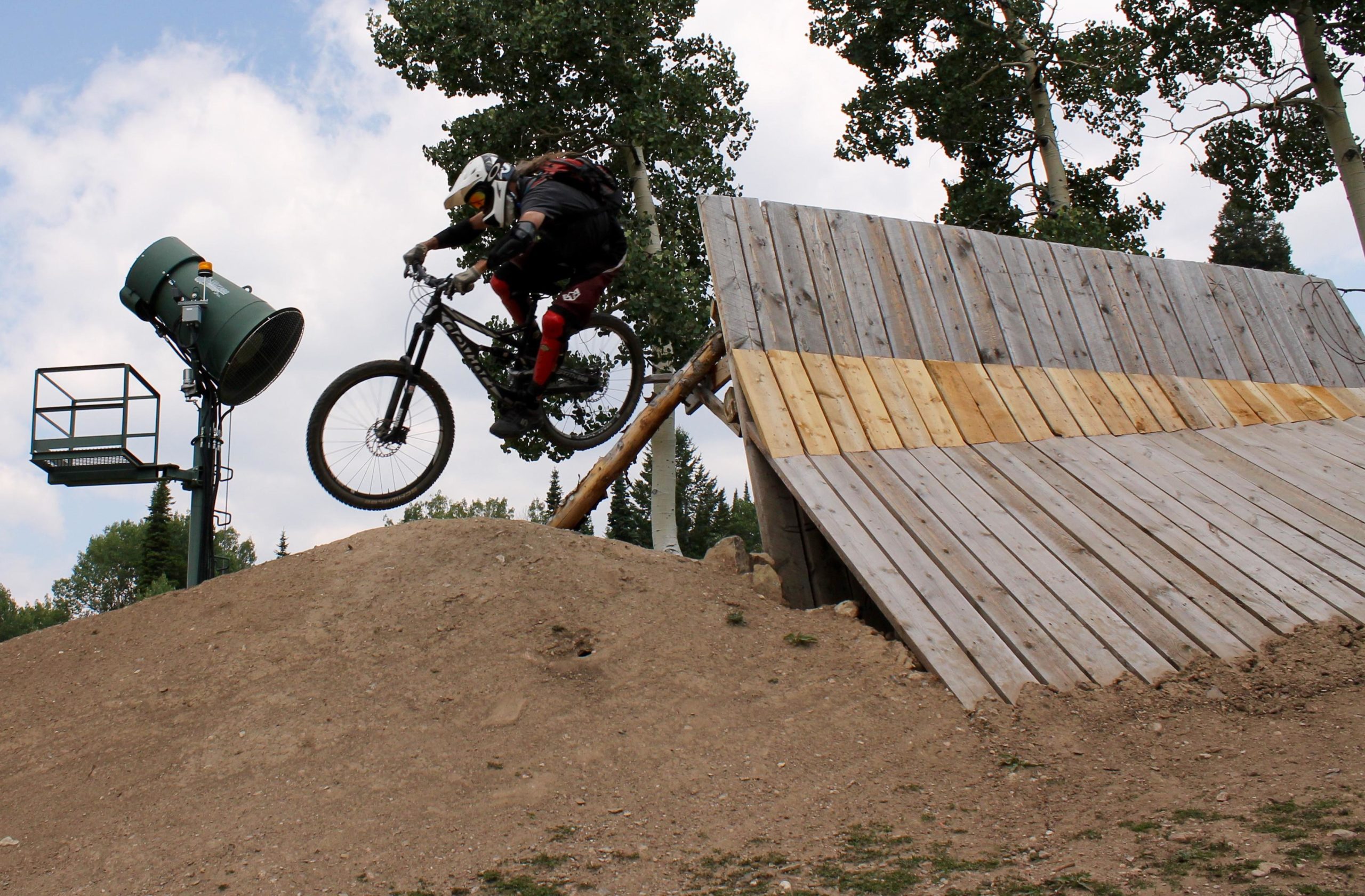 A mountain biker in protective gear jumps off a wooden ramp, airborne above a dirt mound, with trees and a machine in the background on a cloudy day. Grand Targhee Bike Park mountain bike trail.