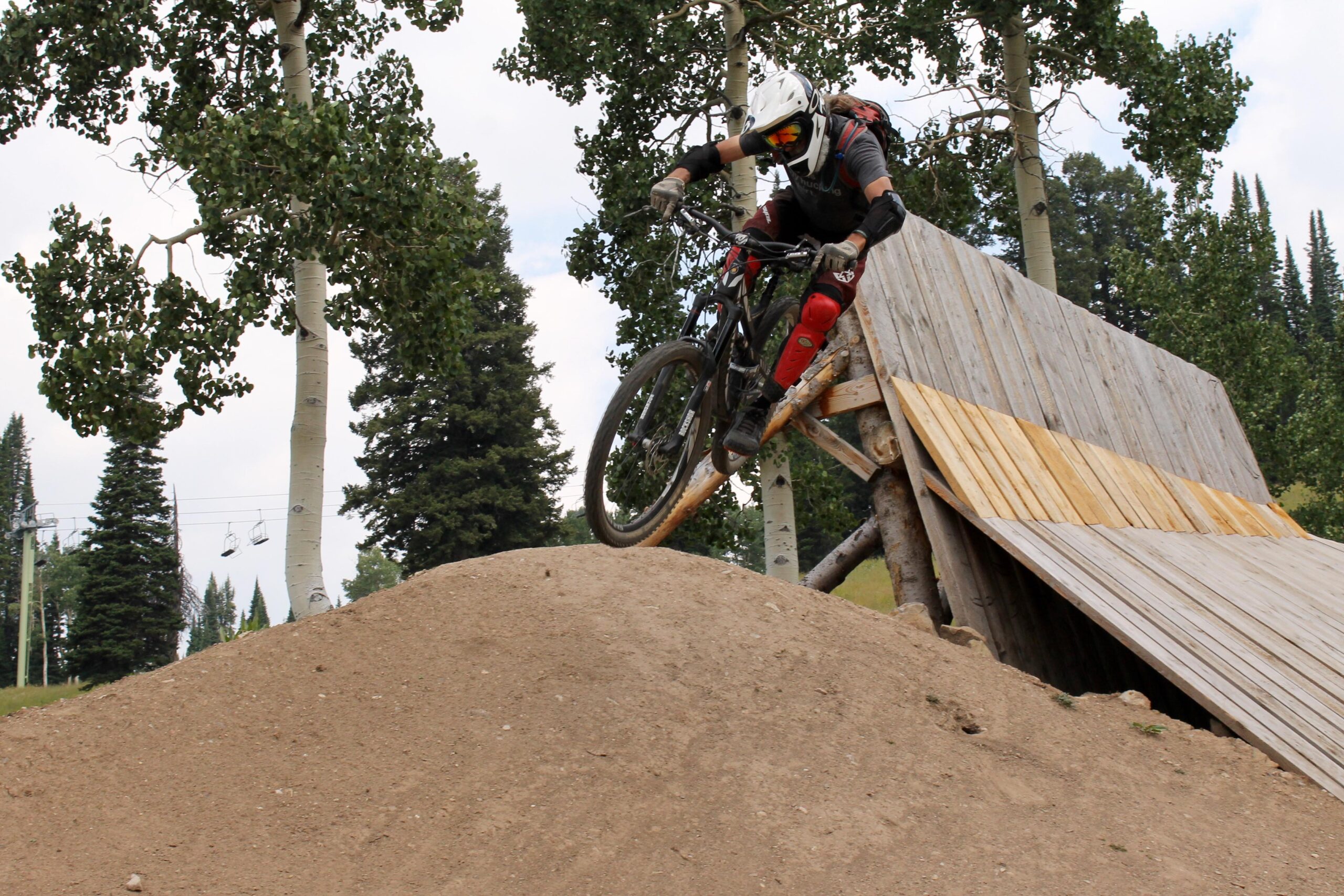 A mountain biker in protective gear jumps off a ramp onto a sandy mound, surrounded by trees and a ski lift in the background. The biker is focused, with a helmet and tinted goggles, showcasing an action-packed moment in a mountainous outdoor setting. Grand Targhee Bike Park mountain bike trail.