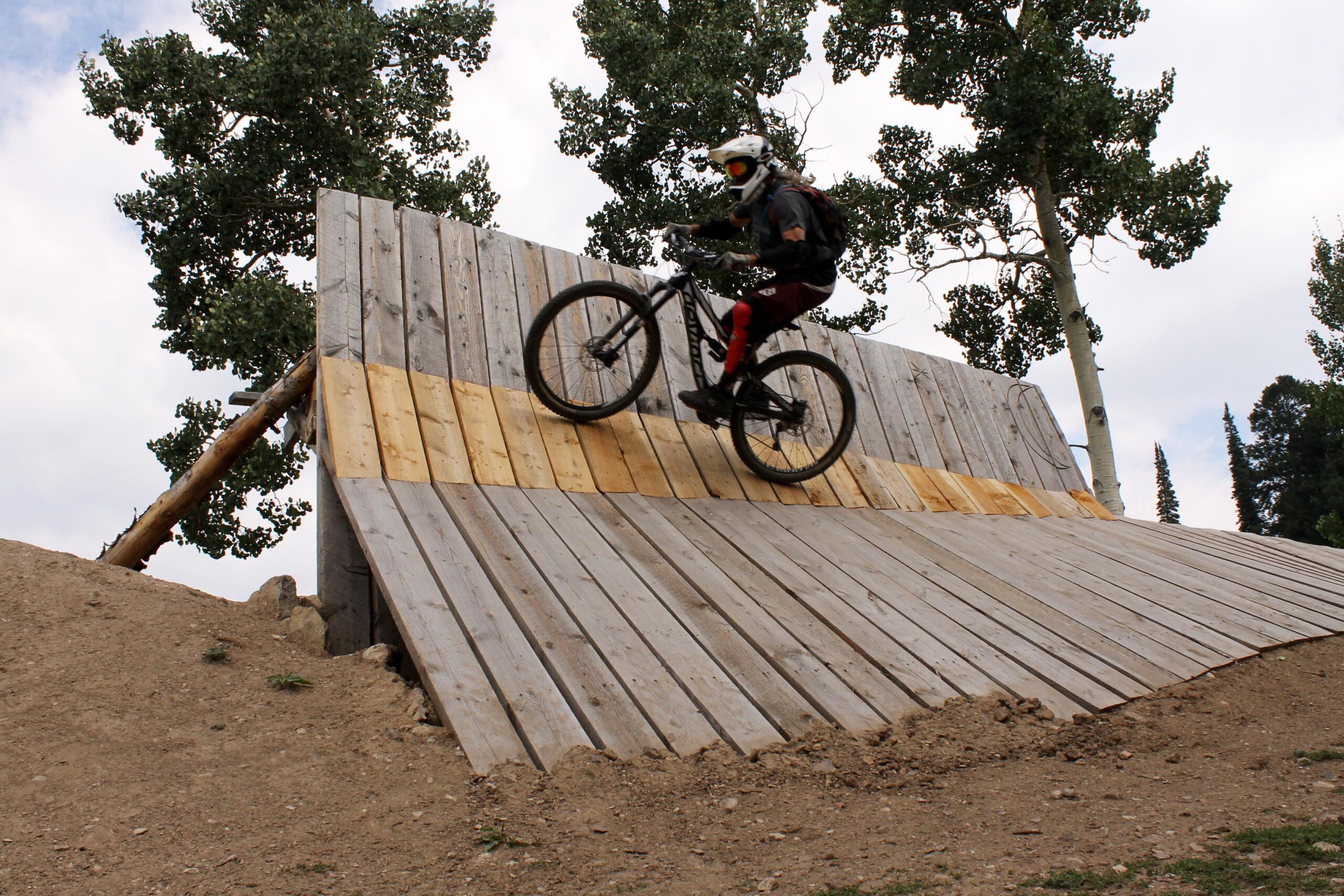 A mountain biker performing a trick on a wooden ramp in a forested area, surrounded by tall green trees under a cloudy sky. Grand Targhee Bike Park mountain bike trail.
