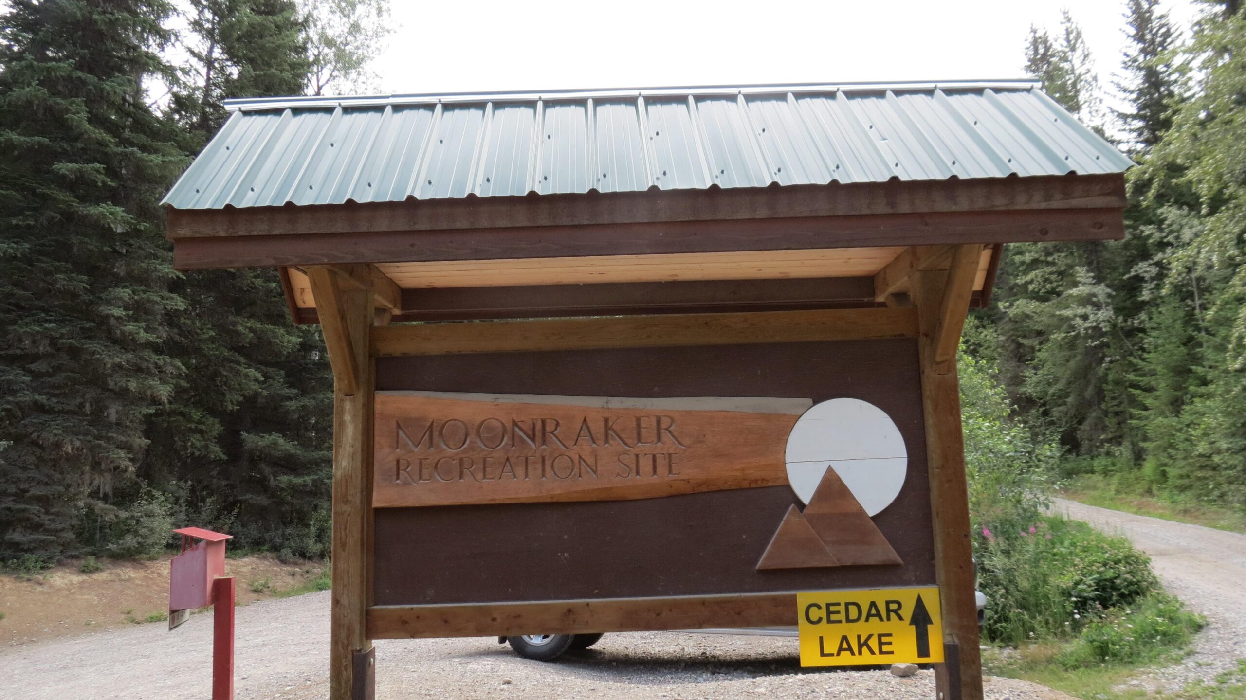 Sign for the Moonraker Recreation Site, featuring rustic wooden design elements, a triangular logo, and directional information pointing towards Cedar Lake, with a backdrop of trees and a gravel road. Moonraker Trails mountain bike trail.