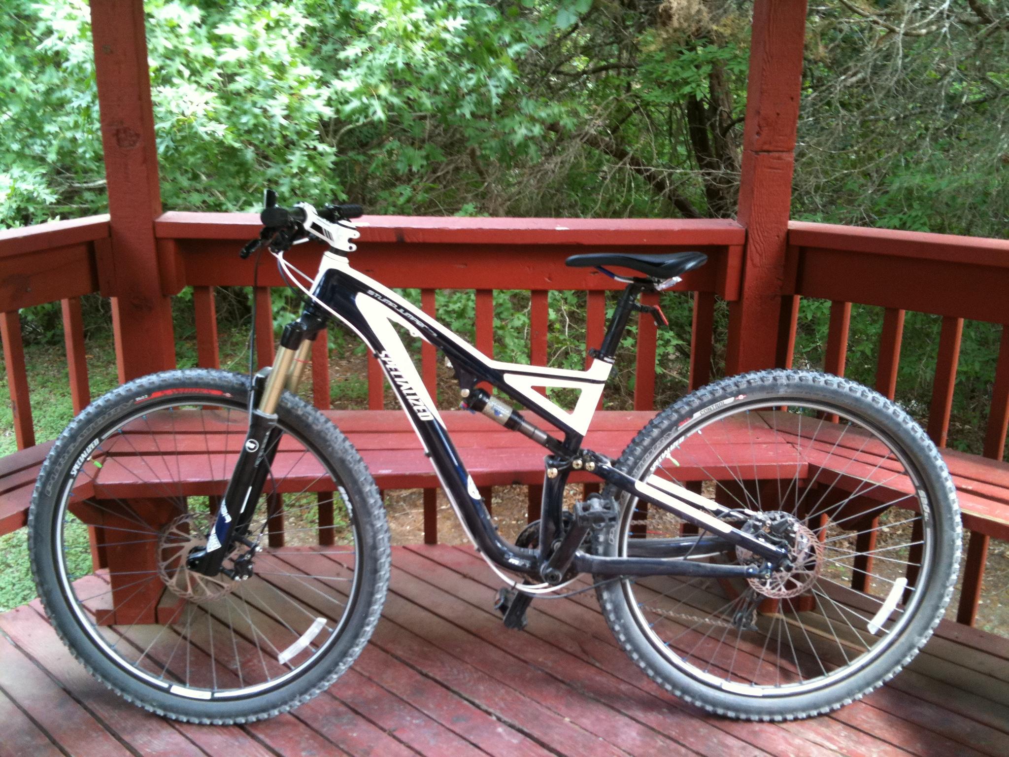Specialized Stumpjumper FSR Comp 29er: A black and white mountain bike positioned on a wooden gazebo deck, surrounded by green foliage. The bike features a front suspension fork and wide, knobby tires, indicating it's designed for off-road cycling.