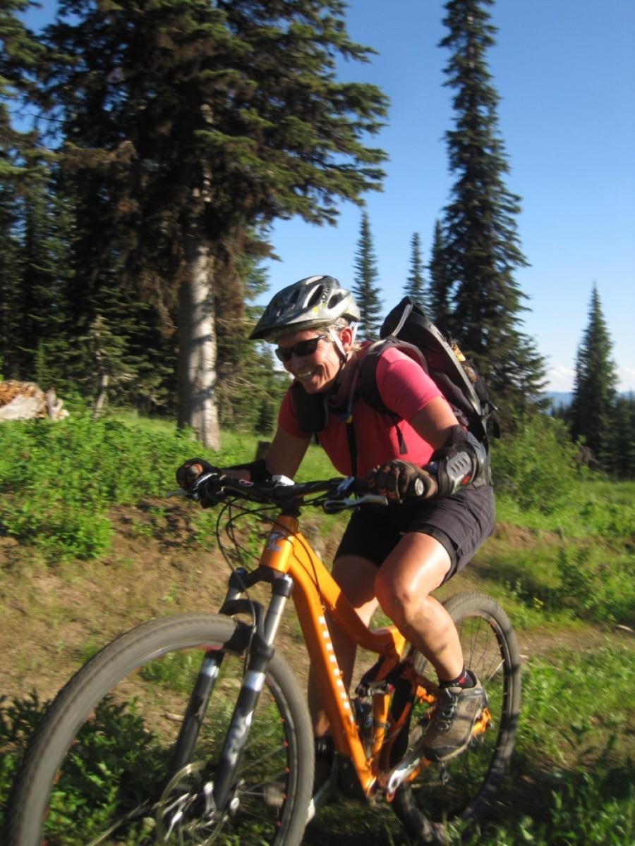 A cyclist in a pink shirt and black shorts is riding an orange mountain bike along a narrow, leafy trail surrounded by tall pine trees. The rider, wearing a helmet and gloves, appears focused and energetic in a sunny outdoor setting. Payette Rim Ride mountain bike trail.