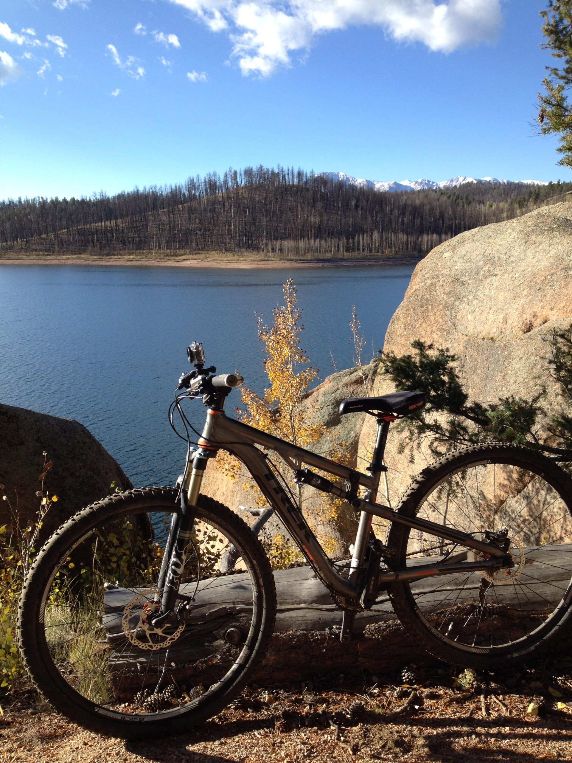 Trek Superfly AL Elite: A mountain bike resting on a log near a calm lake with a backdrop of trees and mountains under a clear blue sky. The scene captures the beauty of nature, highlighting the bike's wheels and frame alongside rocky terrain and autumn foliage.