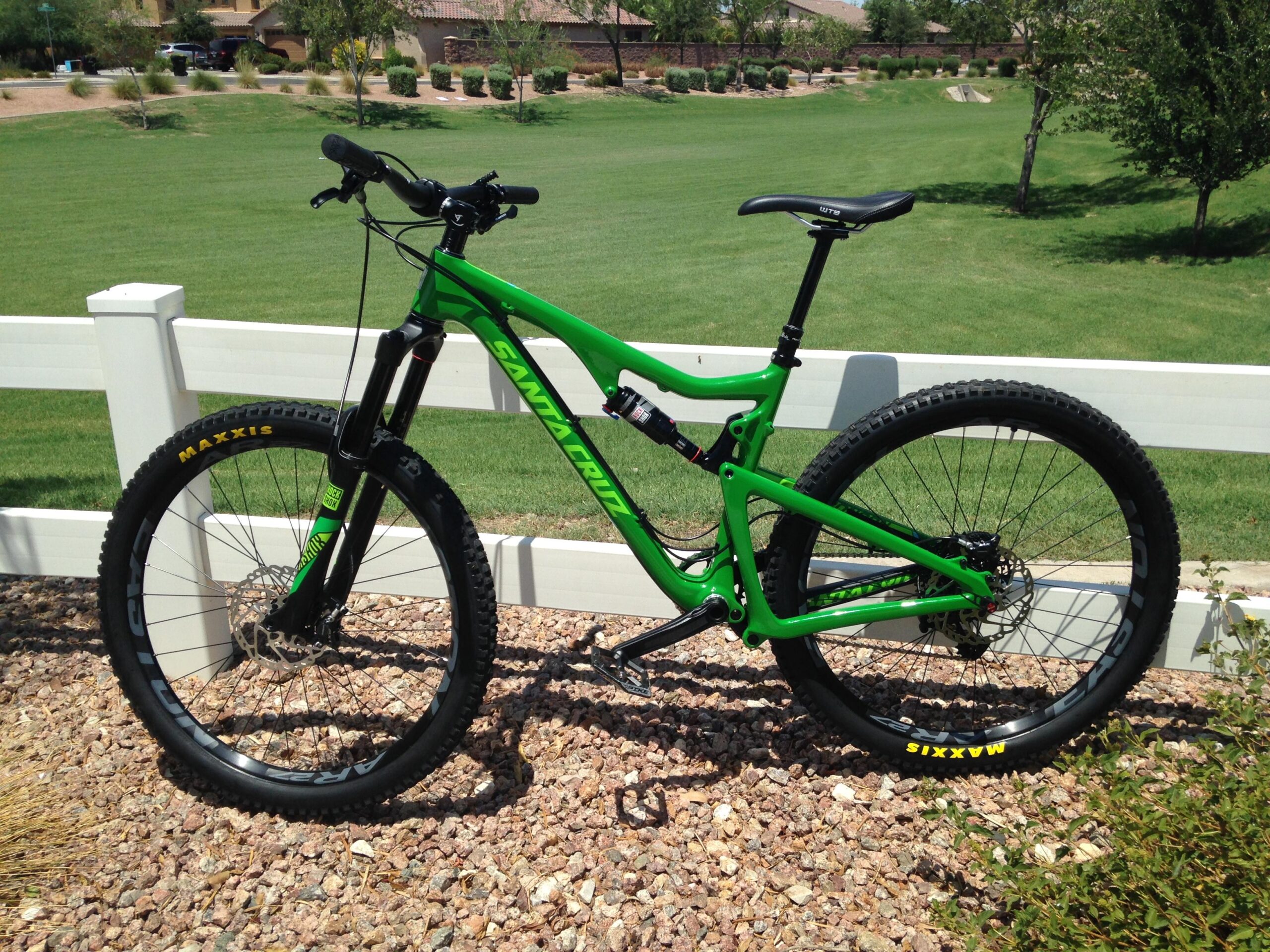 Santa Cruz Bronson: A vibrant green mountain bike positioned against a white fence, set against a grassy background. The bike features thick tires with "Maxxis" branding, a sleek frame design, and is surrounded by pebbles and shrubbery. The scene captures a sunny day with shadow play highlighting the bike's details.