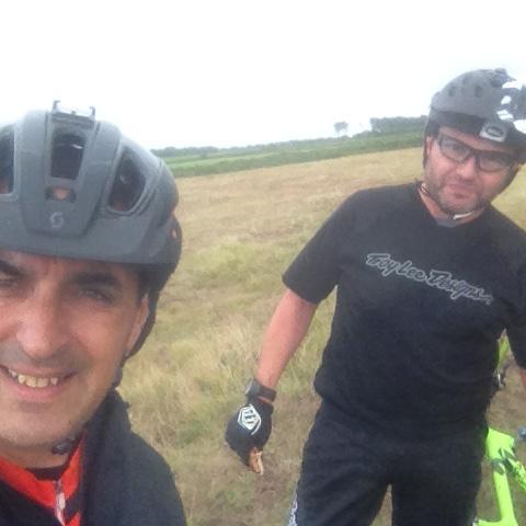 A selfie of two cyclists wearing helmets, standing in a grassy outdoor area. One cyclist is smiling at the camera, while the other stands beside him with a focused expression. The background features a scenic landscape with fields under an overcast sky. Sorel Point to L'Etacq mountain bike trail.