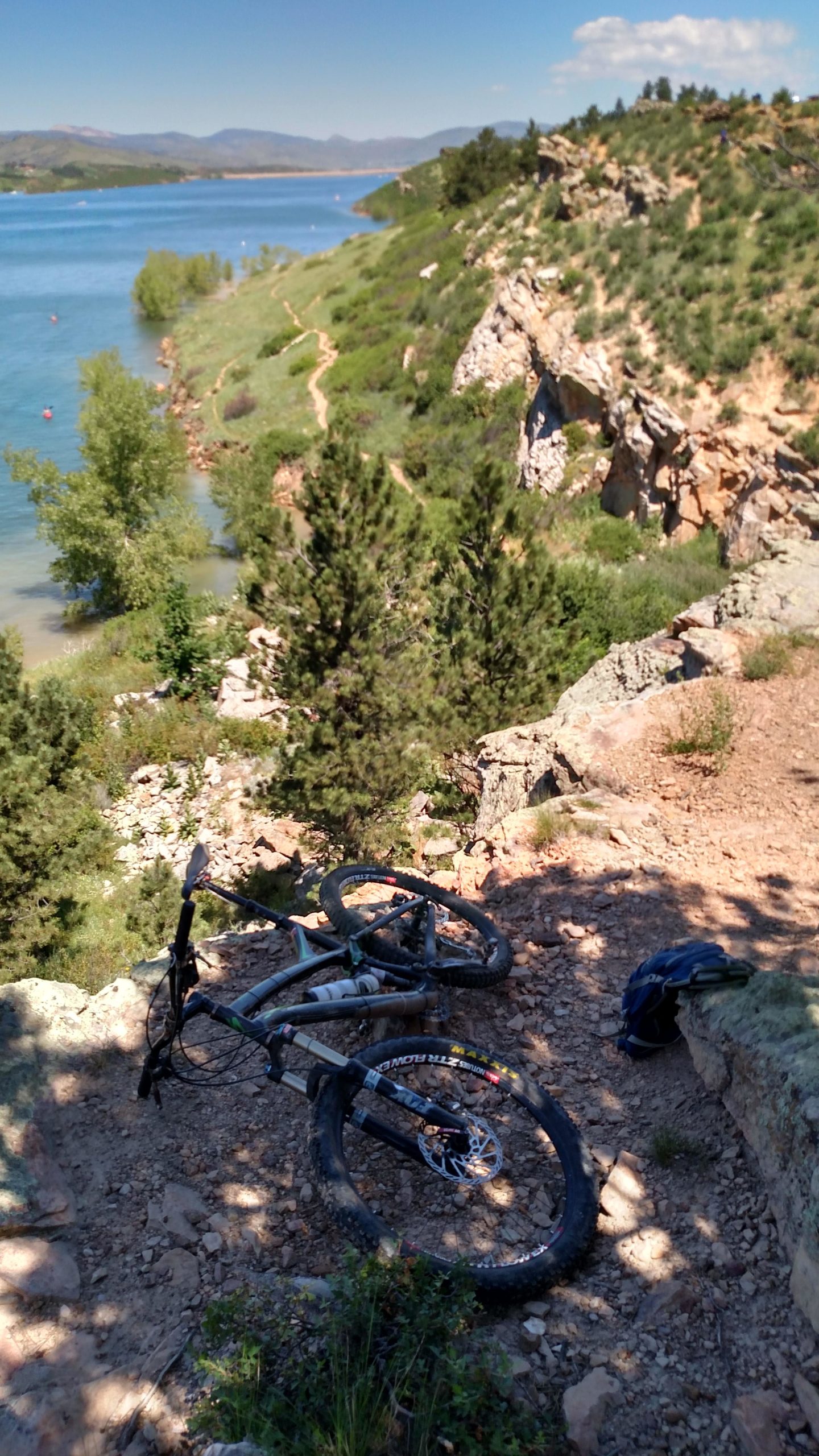 A mountain bike resting on rocky terrain overlooking a serene lake surrounded by greenery and distant mountains under a clear blue sky. Foothills Trail mountain bike trail.