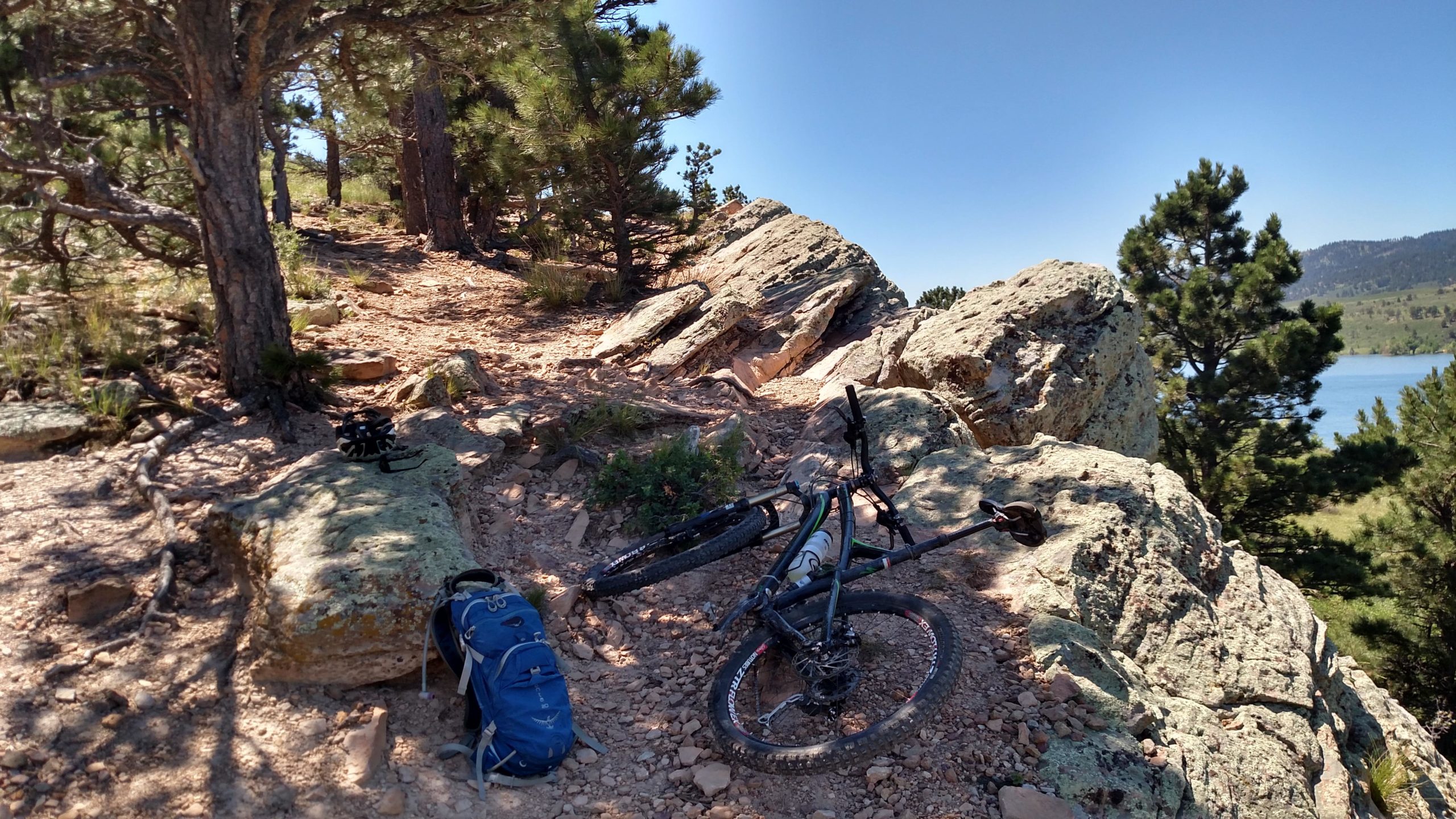 A mountain bike rests on rocky terrain beside a backpack, surrounded by trees and a clear blue sky. In the background, a lake is visible, adding to the scenic outdoor setting. The trail appears rugged, suggesting a hiking or biking adventure. Foothills Trail mountain bike trail.