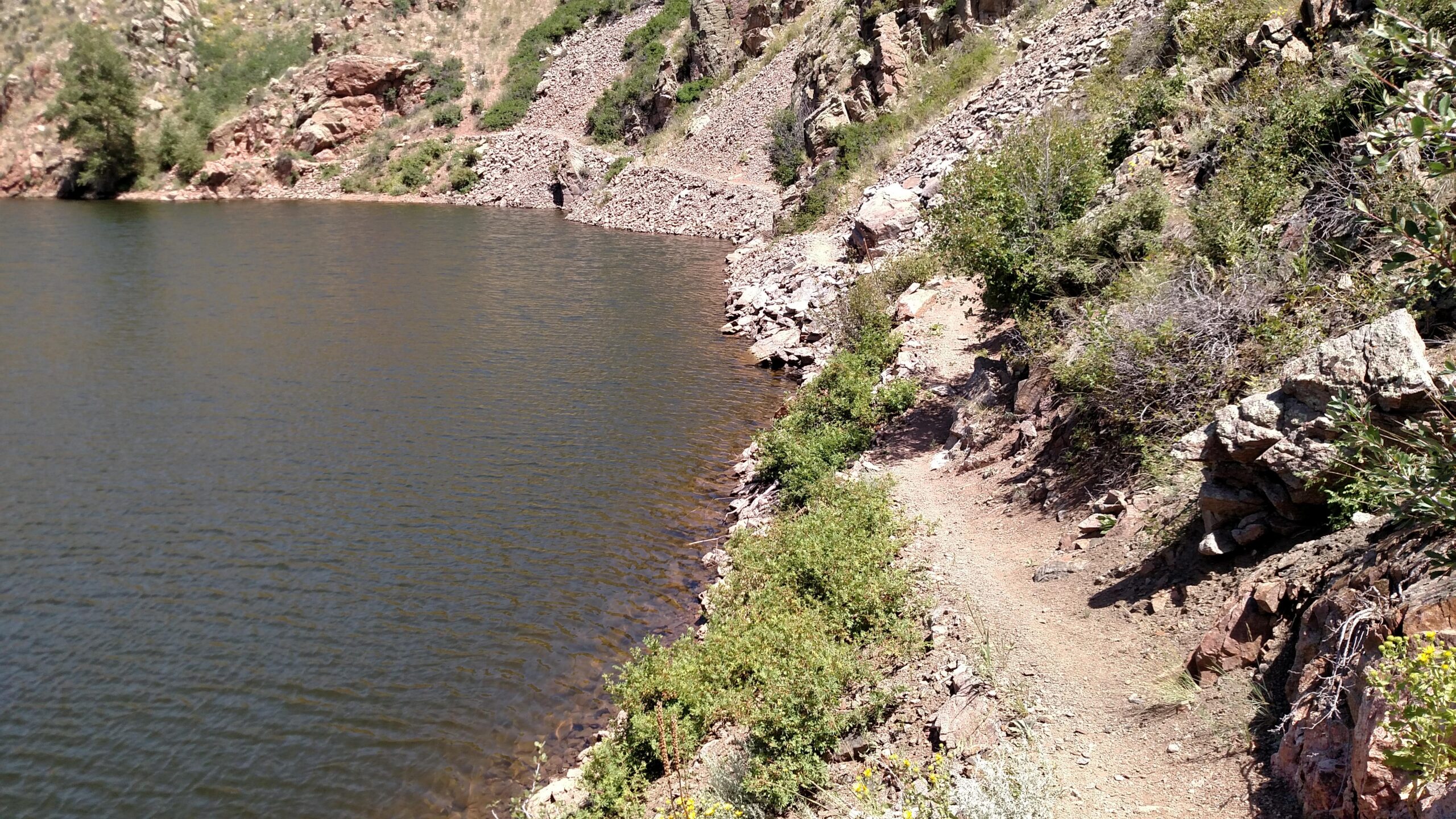 A scenic view of a rocky shoreline with gentle waves lapping against the water's edge, surrounded by lush greenery and steep hills. A narrow, winding path runs along the edge, inviting exploration of the natural landscape. Canyons mountain bike trail.