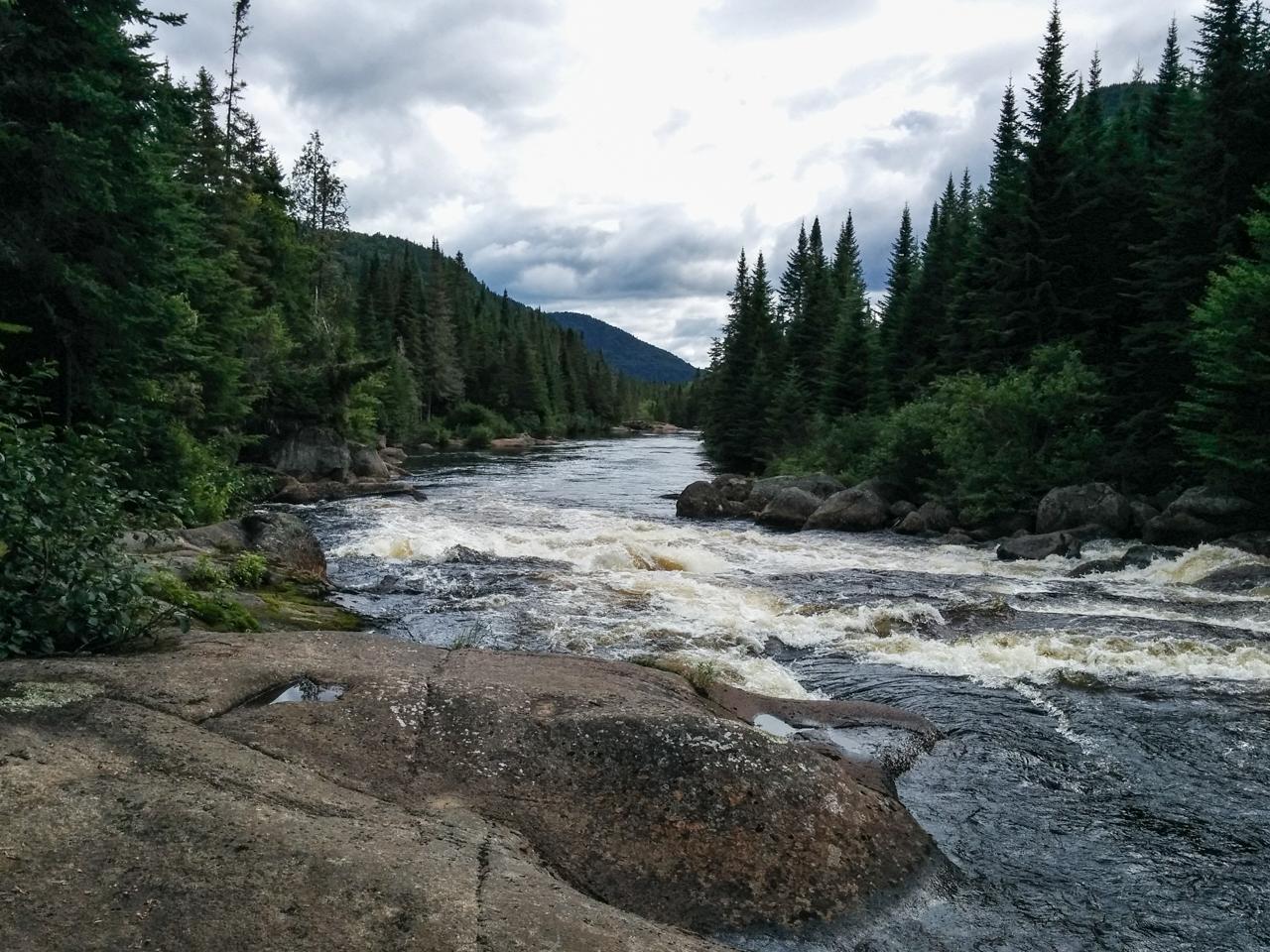 A scenic view of a river flowing through a forested landscape, with rocky banks and lush greenery on both sides. The sky is partly cloudy, and distant mountains can be seen in the background. The water appears to be rushing, creating small waves and white foam. Vallee Bras Du Nord Secteur Shannahan mountain bike trail.