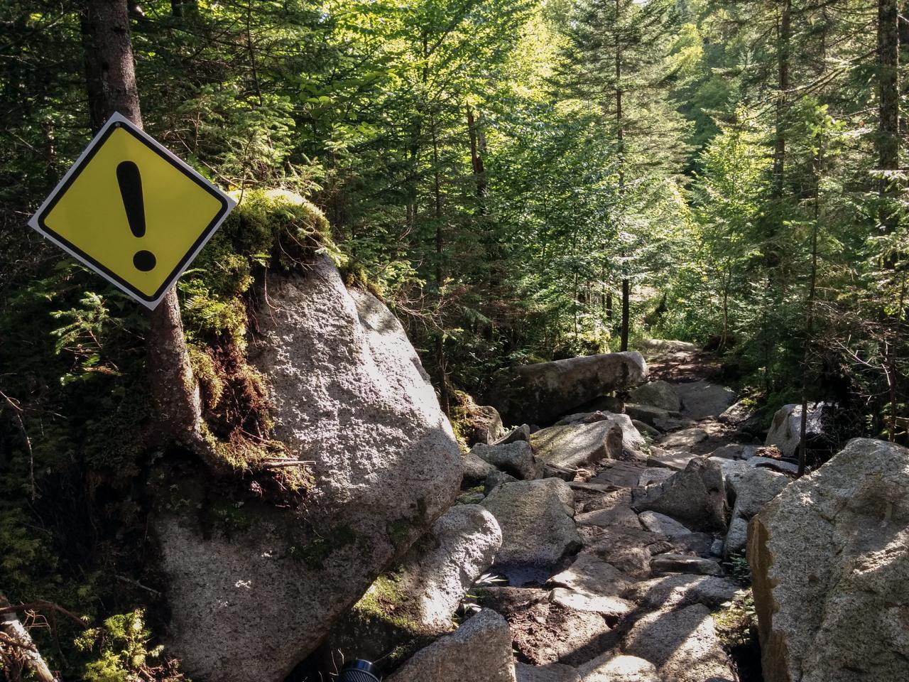 A rocky hiking trail surrounded by trees, with a yellow caution sign featuring an exclamation mark mounted on a tree near the path. The area is well-lit by sunlight filtering through the leaves. Vallee Bras Du Nord Secteur Shannahan mountain bike trail.