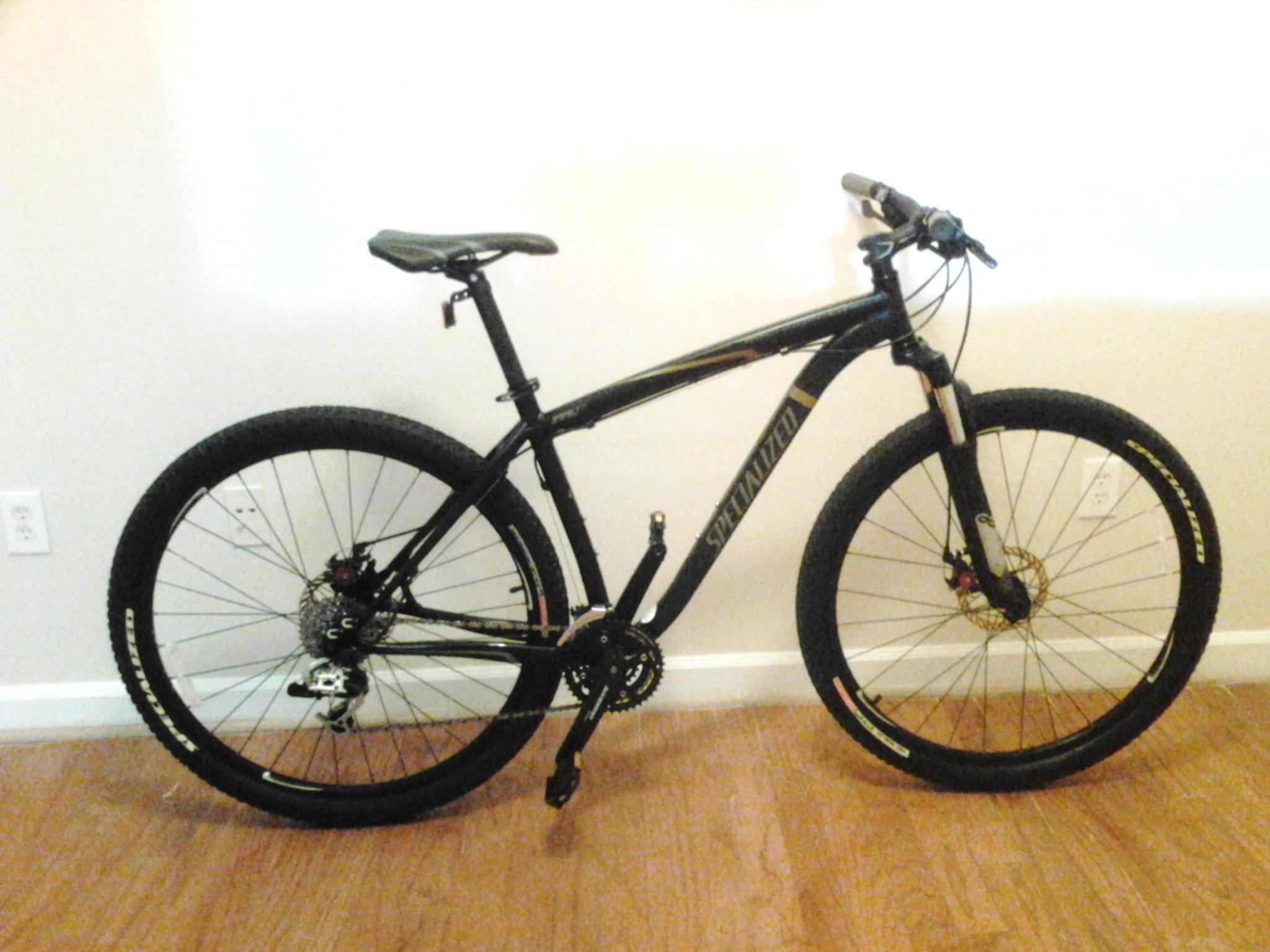 Specialized Rockhopper Expert Disc 29er: A black mountain bike with thick tires and a sleek frame, positioned against a light wall. The bike features disc brakes, a detailed gear system, and a comfortable seat, with hardwood flooring visible in the background.