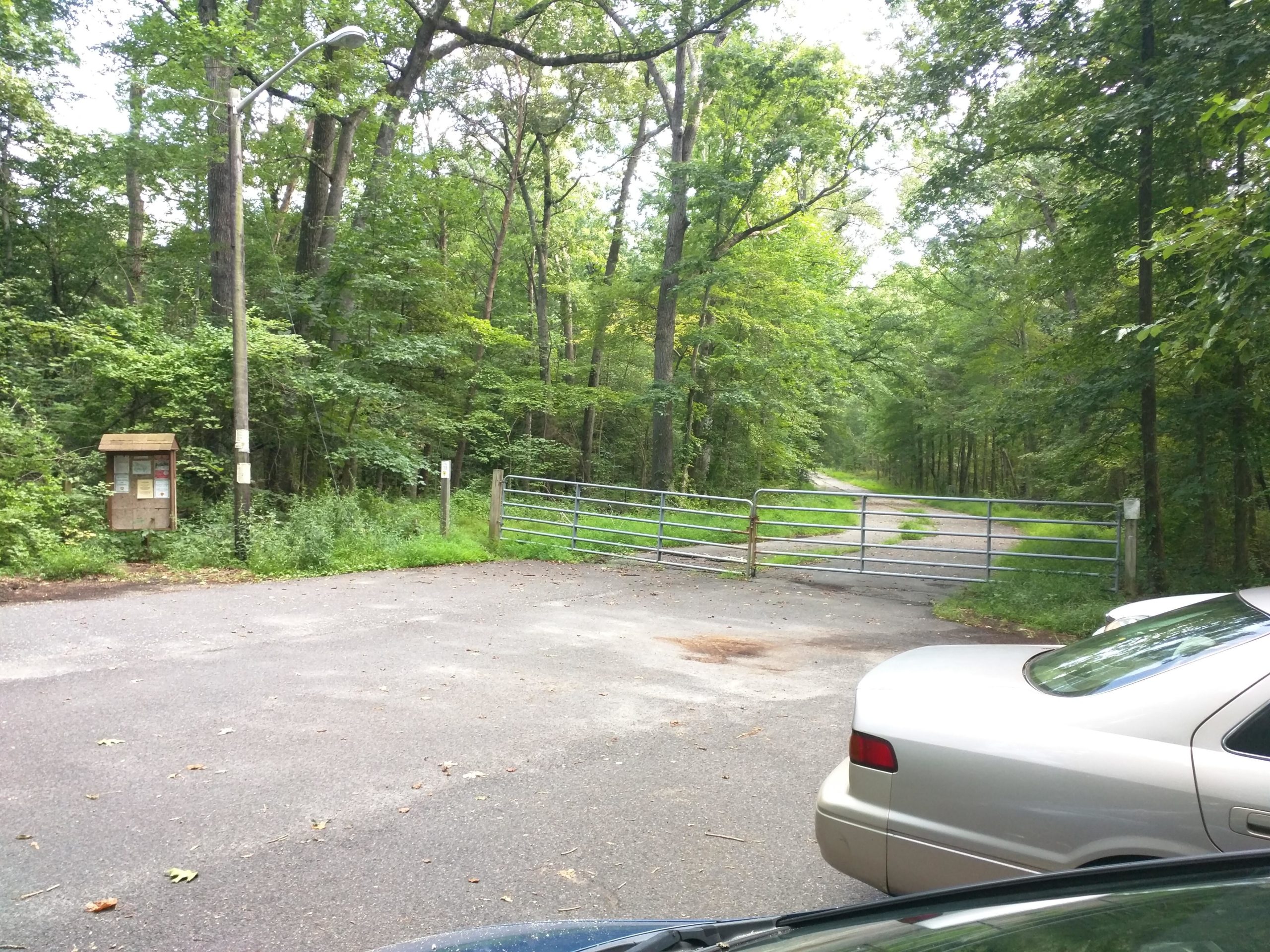 A gravel road leading into a wooded area is partially blocked by a metal gate. To the left, there is a wooden information kiosk with posted notices. Nearby, a streetlight stands alongside the road, which is lined with trees. Two parked cars are visible in the foreground. The scene is set in a natural, green landscape suggesting an entrance to a park or nature reserve. Rancocas State Park - Westampton mountain bike trail.