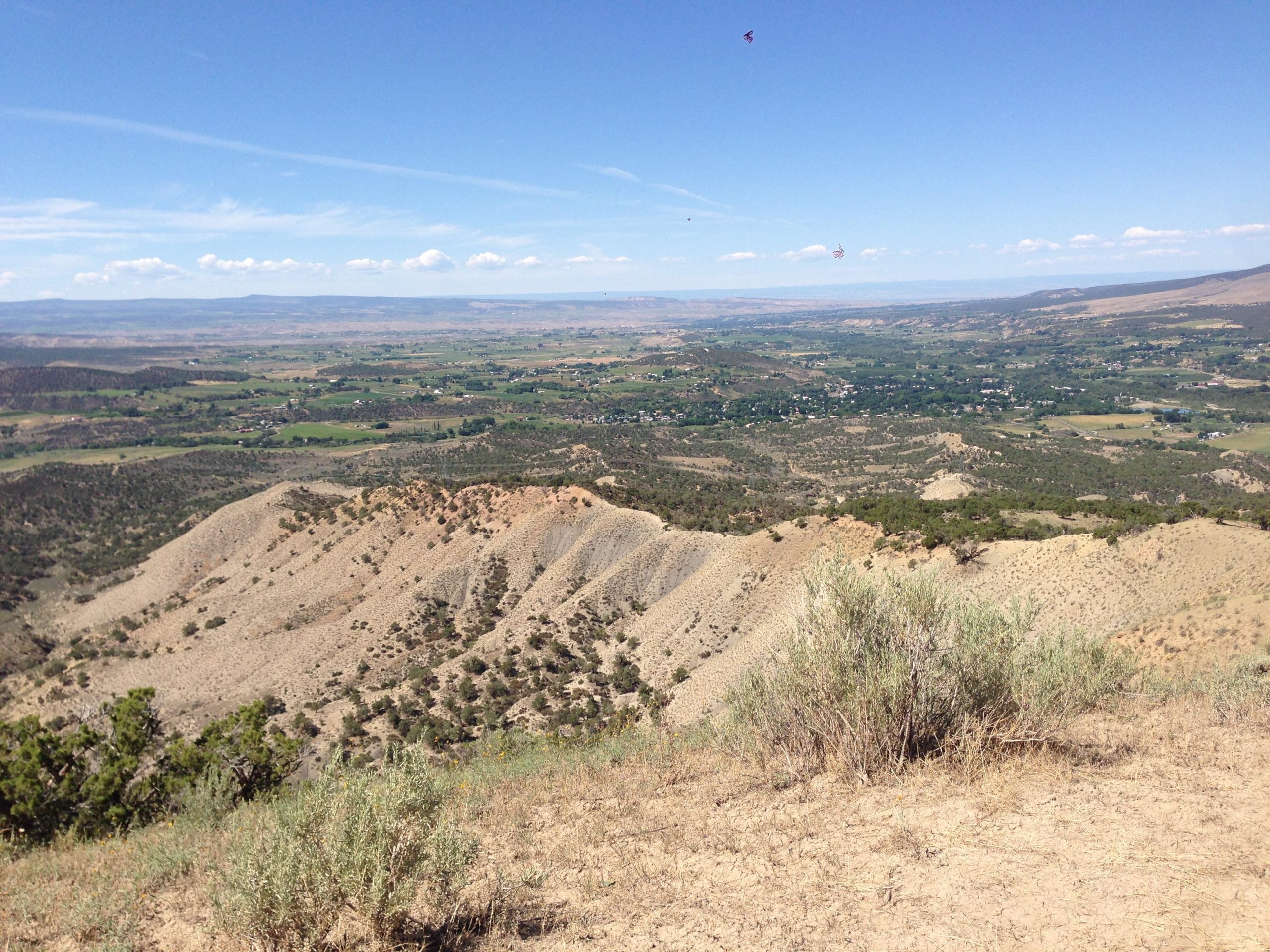 A panoramic view of a mountainous landscape with rolling hills, scattered vegetation, and patches of farmland below, set against a bright blue sky with scattered clouds. The foreground features dry, sandy terrain with sparse shrubs. Jumbo Mountain mountain bike trail.