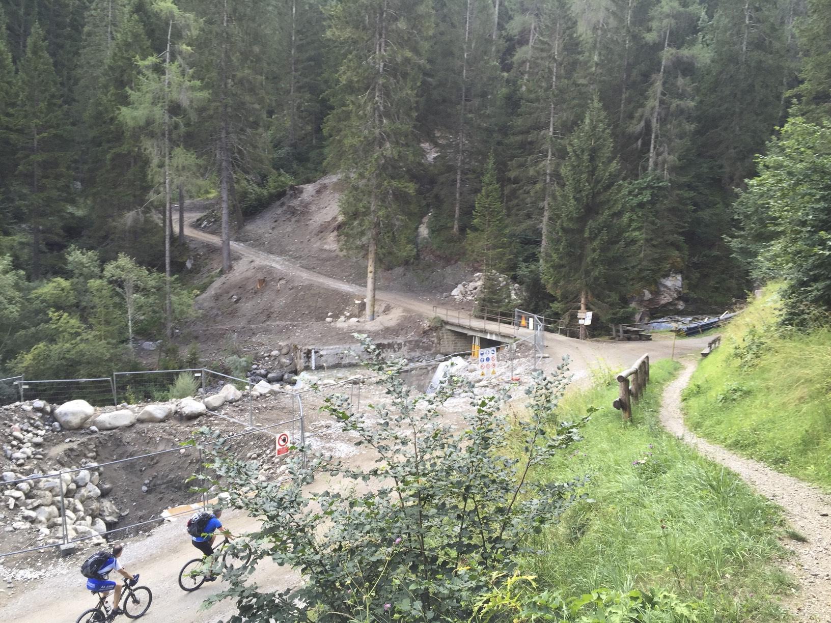 Two mountain bikers ride along a dirt path in a forested area, with trees lining the route and a rocky construction site visible in the background. A wooden bridge and various signs are also present along the path. The scenery is lush and green, indicating a natural outdoor environment. Campiglio-Dimaro mountain bike trail.