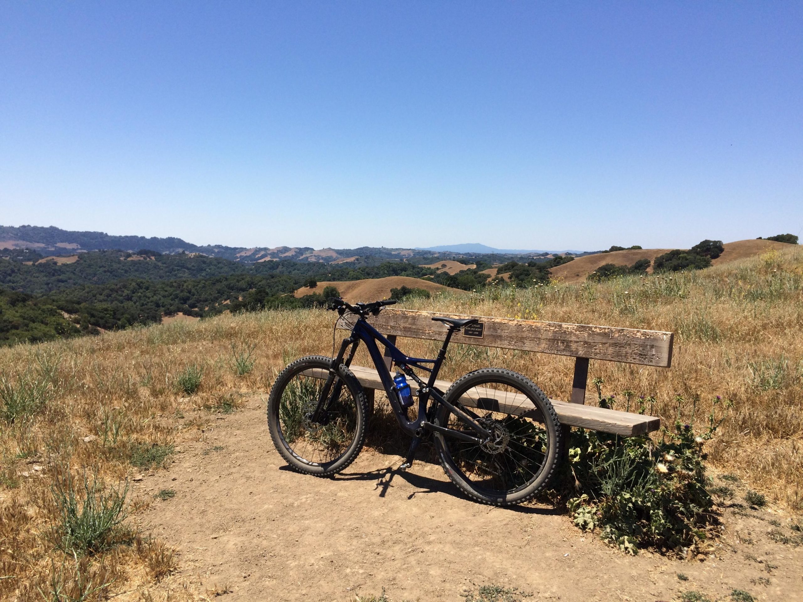 A blue mountain bike leaned against a wooden bench overlooking a scenic view of rolling hills and greenery under a clear blue sky. Briones Regional Park mountain bike trail.