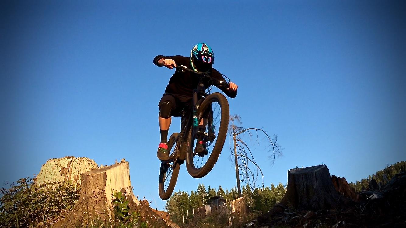 A person in a black outfit and helmet is performing a jump on a mountain bike over a tree stump, with a clear blue sky and forest in the background. The scene captures the thrill of mountain biking in a natural setting. Cold Creek mountain bike trail.