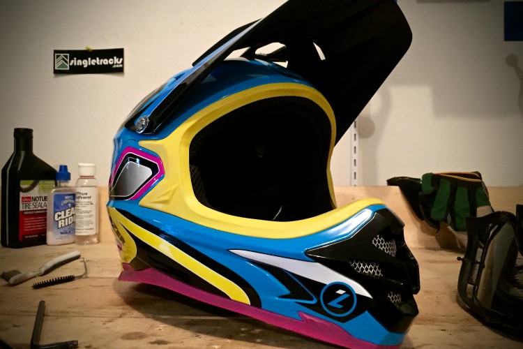 A brightly colored motocross helmet in blue, yellow, and pink sits on a wooden work surface. Various maintenance supplies, including oils and cleaning products, are arranged nearby, along with an allen wrench and a pair of gloves. A sticker on the wall in the background features the logo of a biking website.